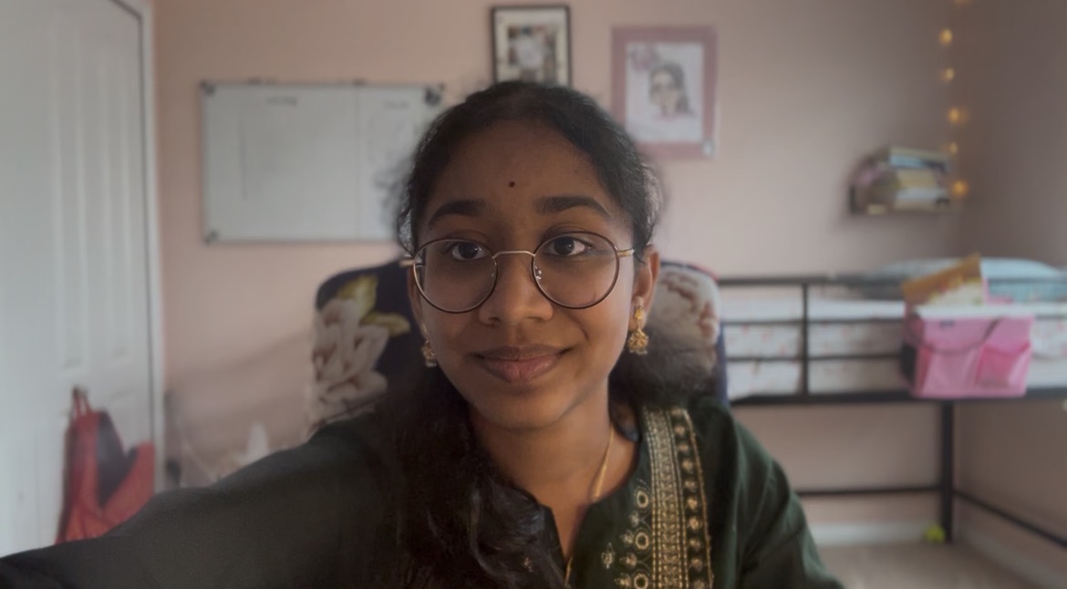 Young girl with glasses smiling in a room with a bed and shelves in the background.