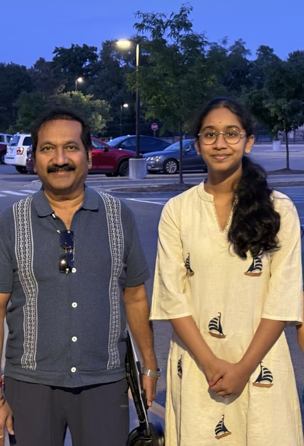 Two people standing outdoors in front of parked cars and trees, smiling, with streetlights in the background.
