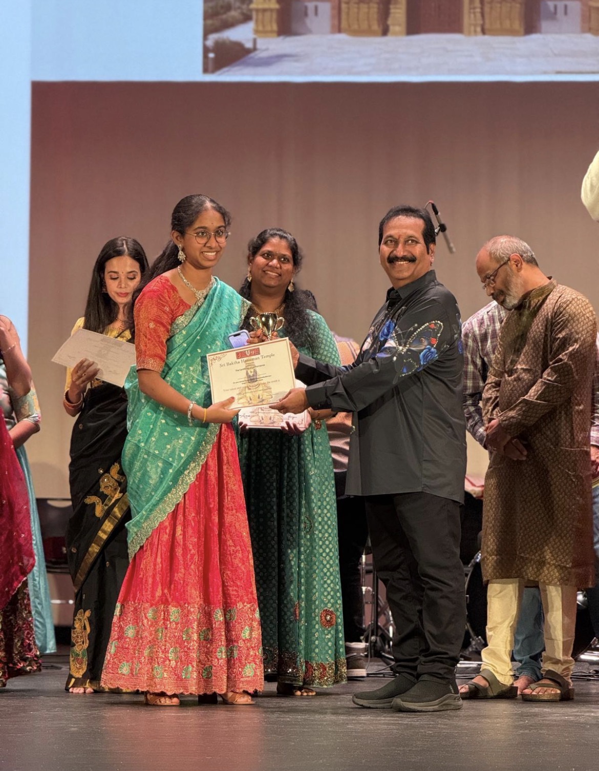 Group of people on stage, woman in green and red saree receiving certificate from man in black shirt, others watching.