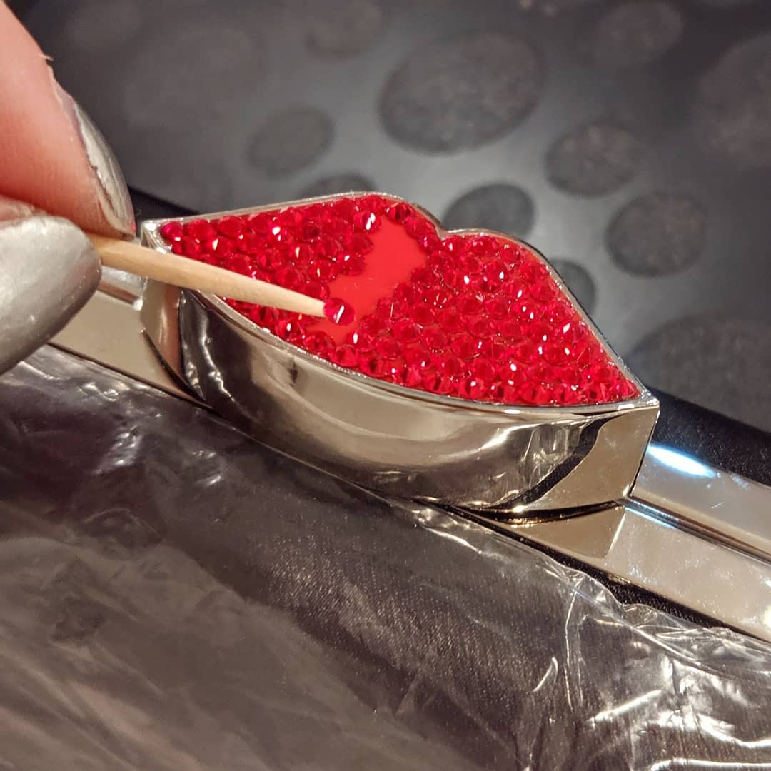A heart-shaped metal mold filled with red sugar crystals, being decorated with a toothpick, on a dark surface.
