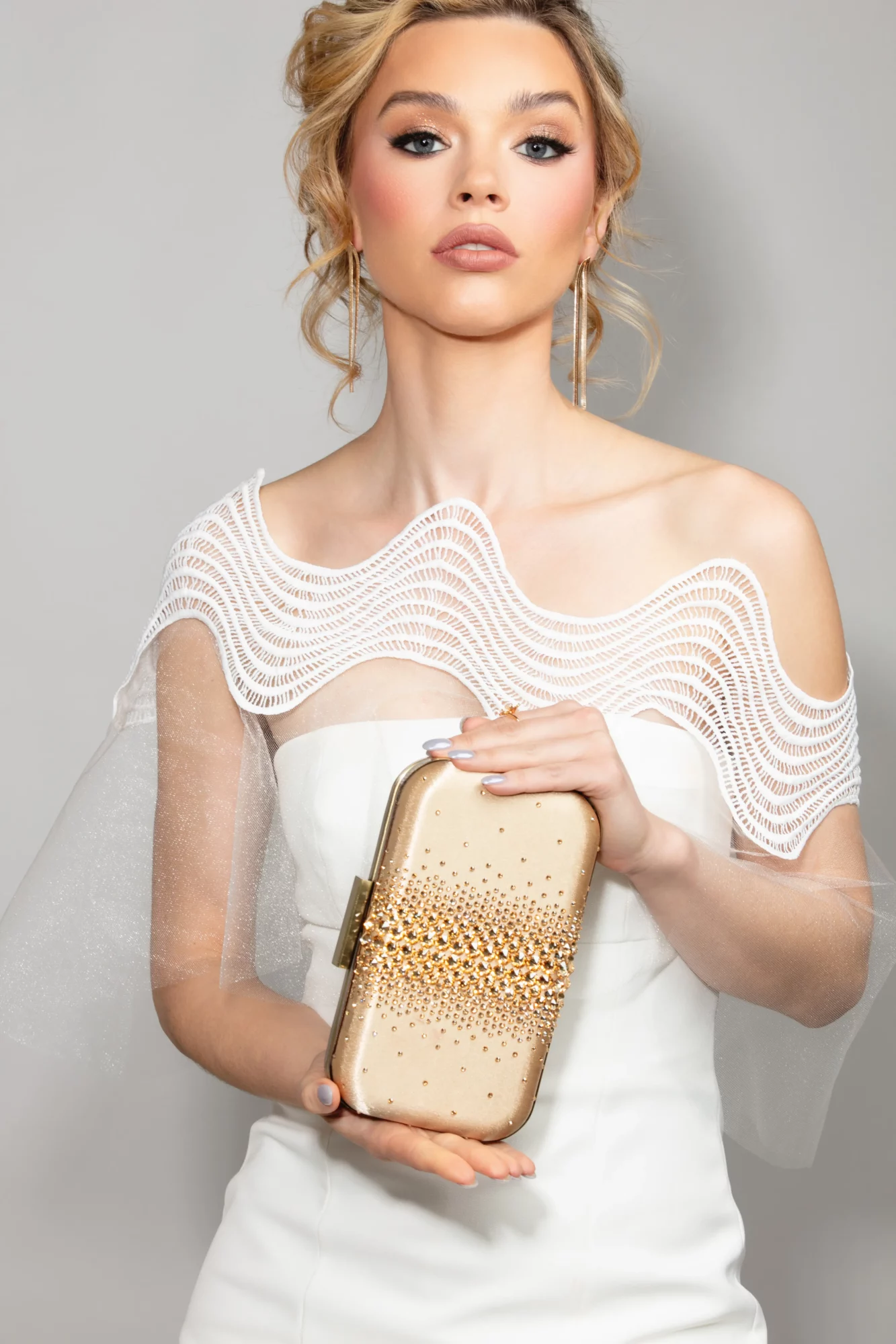 Young woman with blonde hair in an elegant white dress holding a gold clutch bag, standing against a plain background.