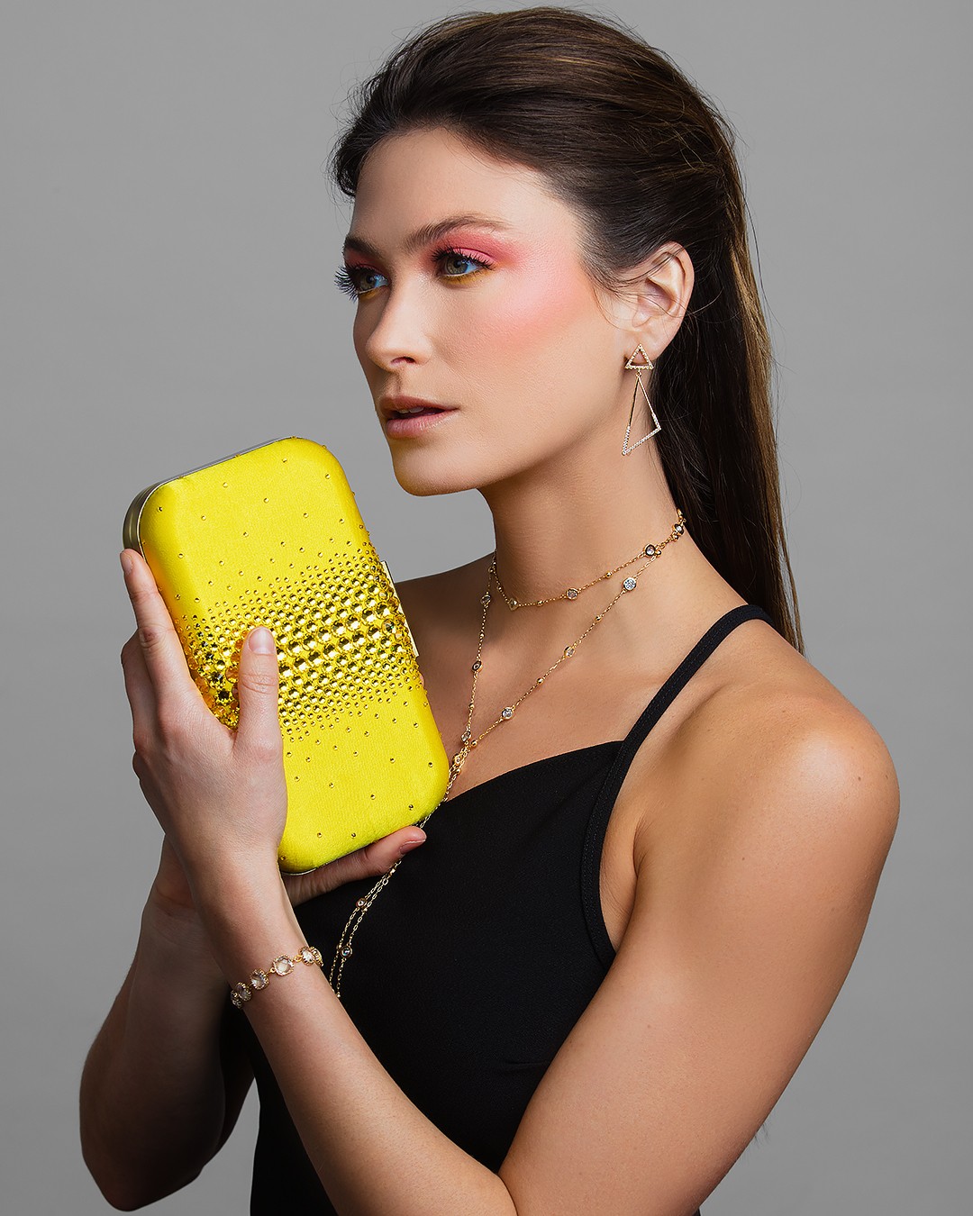 Woman with dark hair, wearing a black top and jewelry, holding a yellow clutch bag, against a gray background.