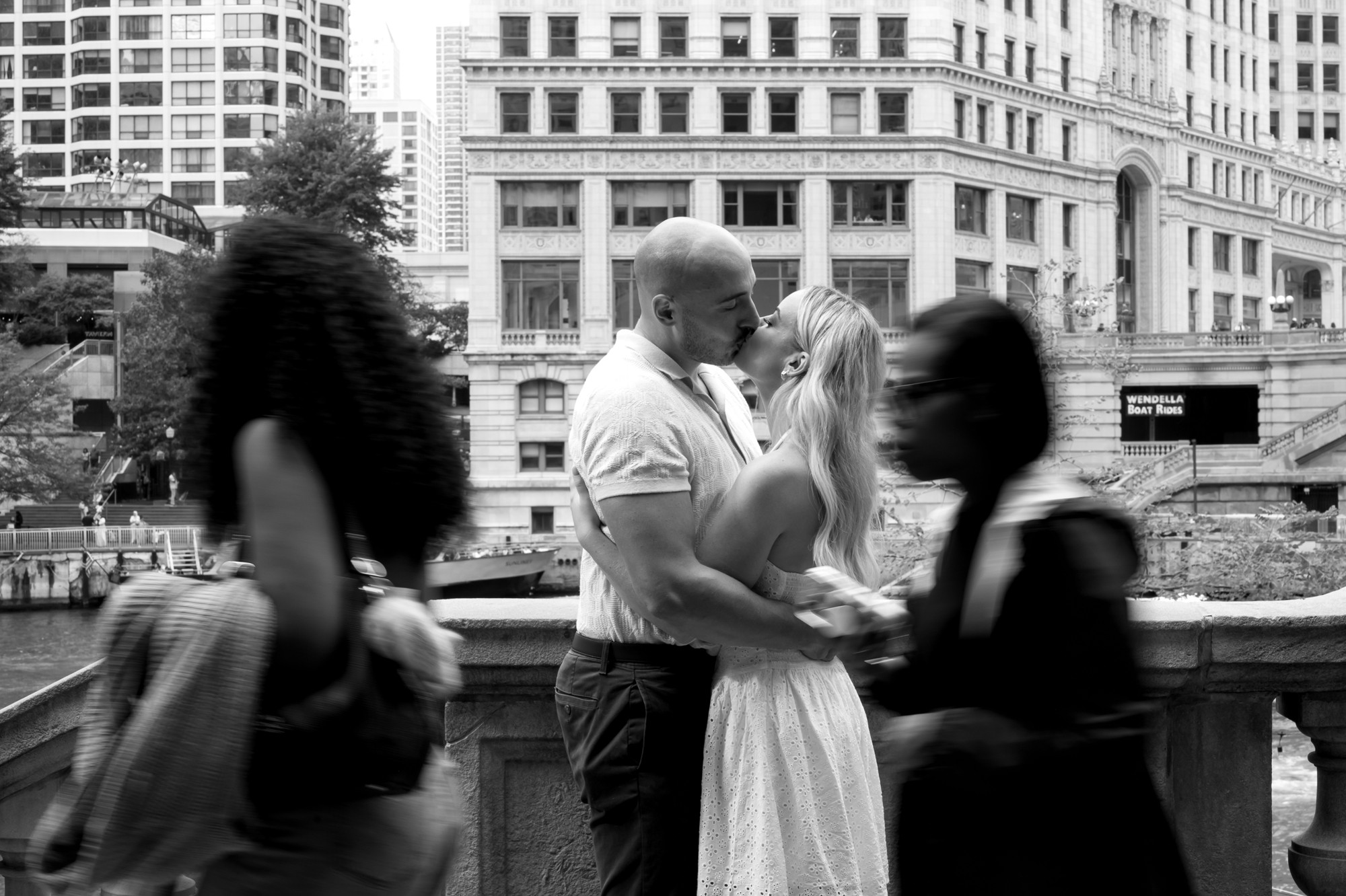 A couple kissing outdoors with city buildings in the background, surrounded by blurred people, in black and white.