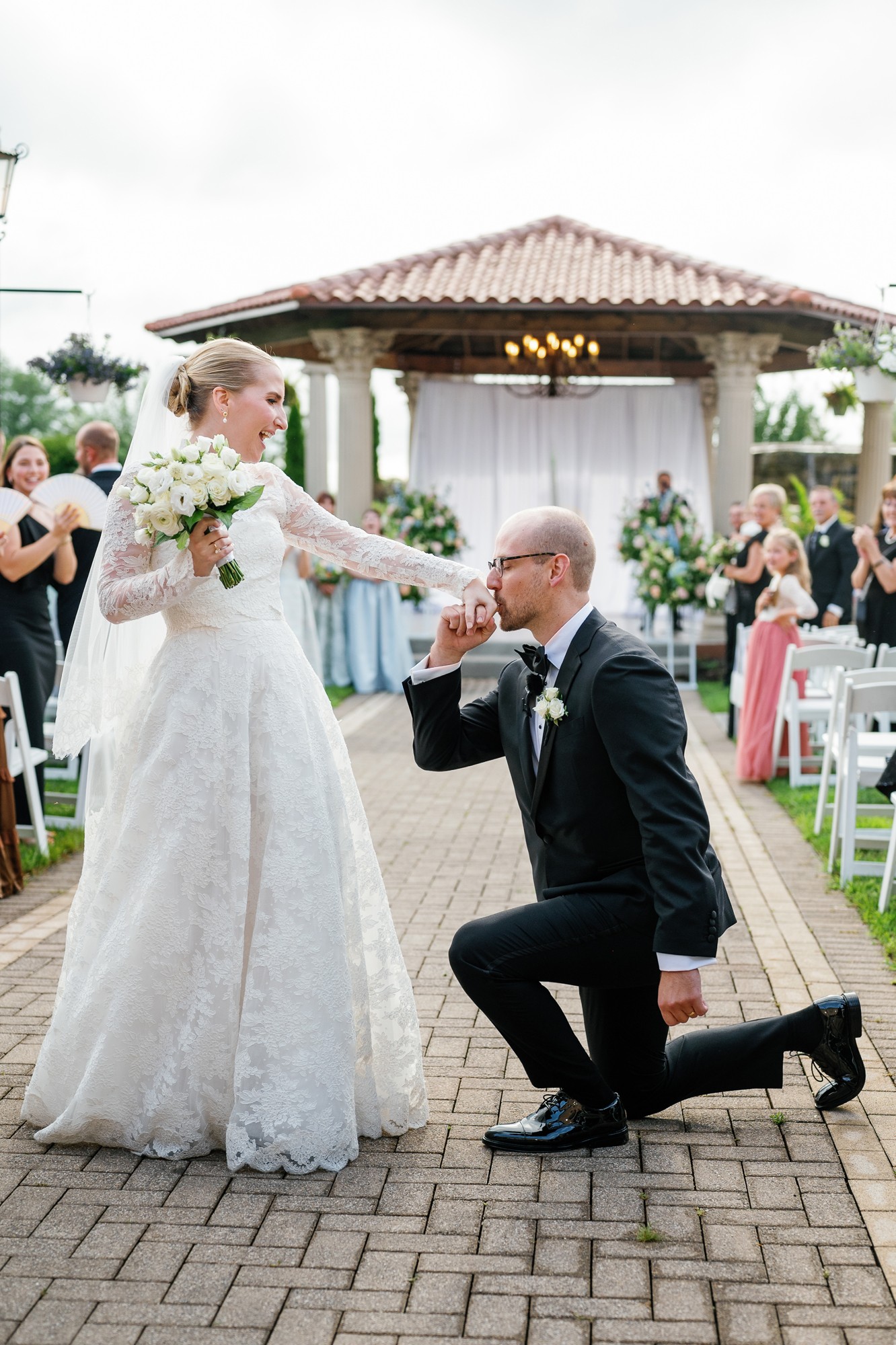 Groom kneeling and proposing to bride holding a bouquet outdoors during wedding ceremony, with guests and a gazebo in background.
