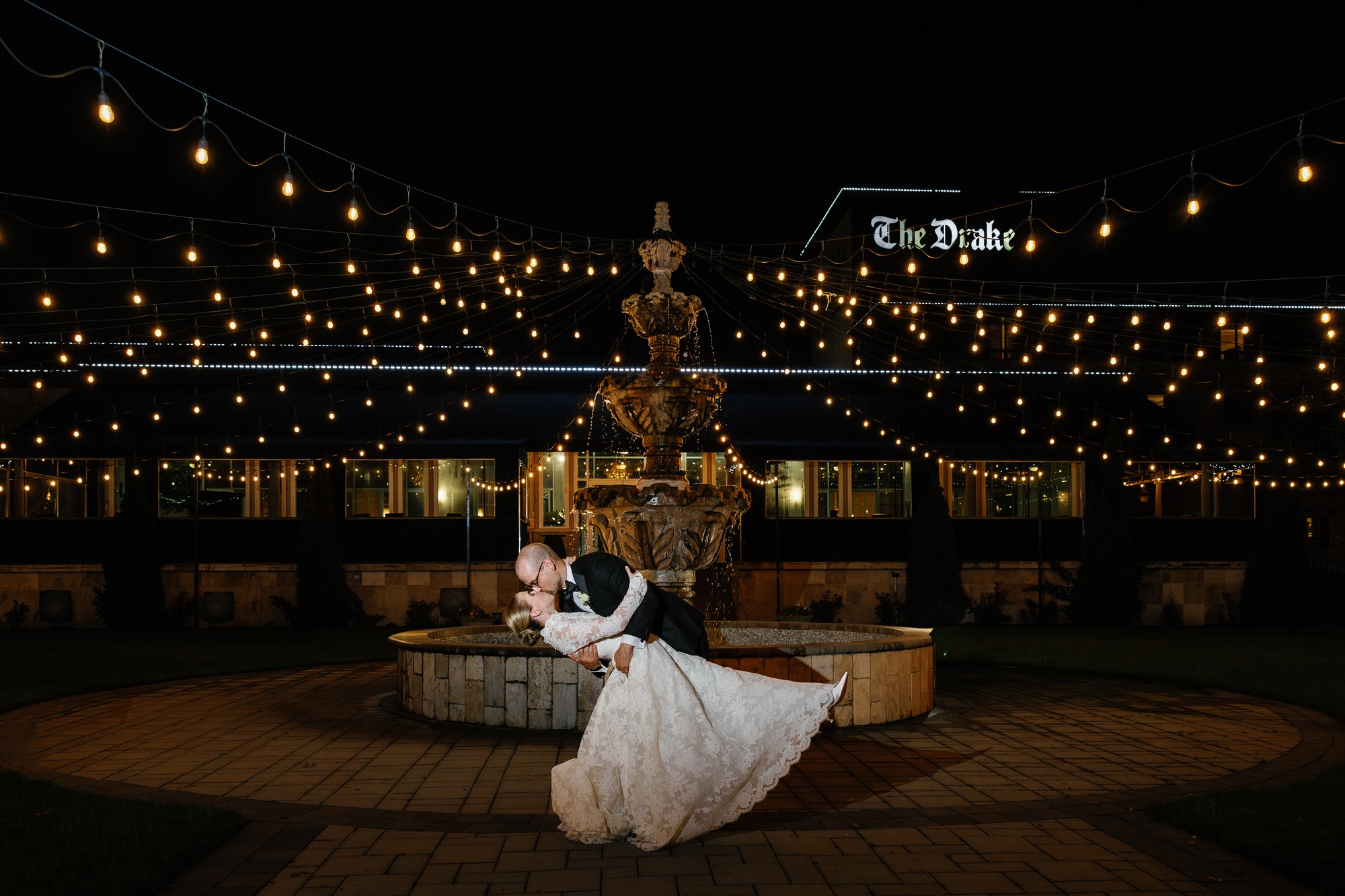 A bride and groom dance in front of a fountain illuminated by string lights at night, with a building and sign in the background.