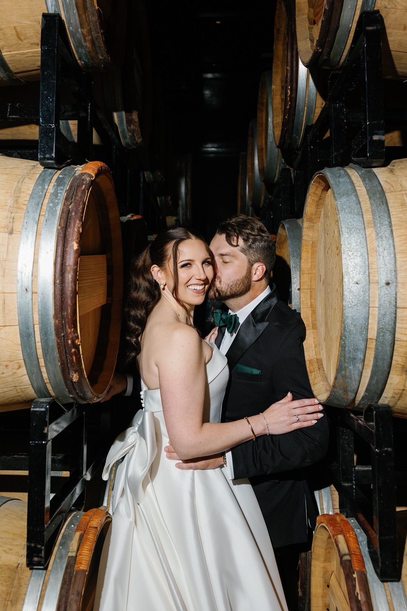 A bride and groom in wedding attire embrace and smile among stacked wine barrels.