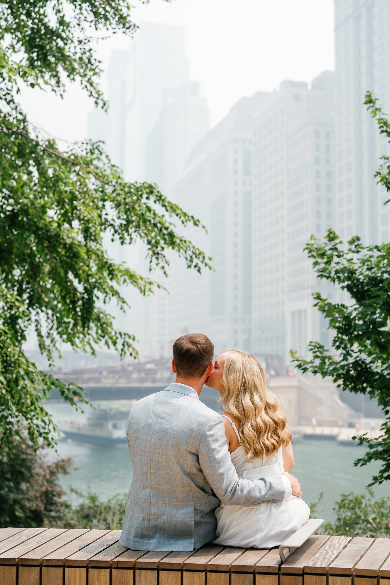 A couple sits on a wooden platform overlooking a river with tall city buildings in the background, surrounded by green trees.