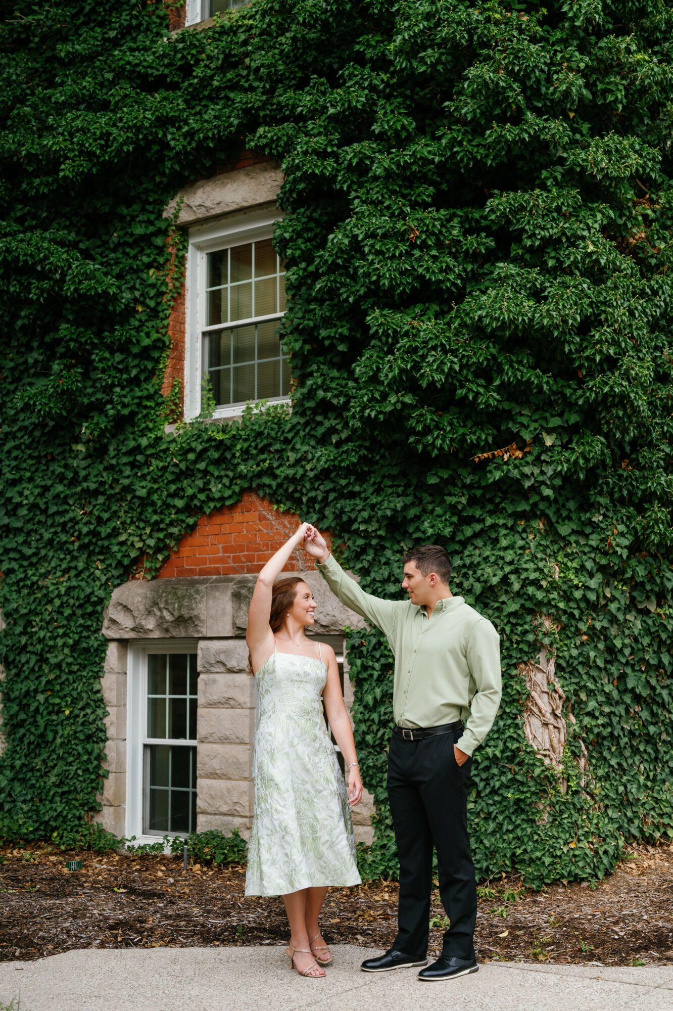 A couple stands outside a brick building covered in green ivy, holding hands and looking at each other.