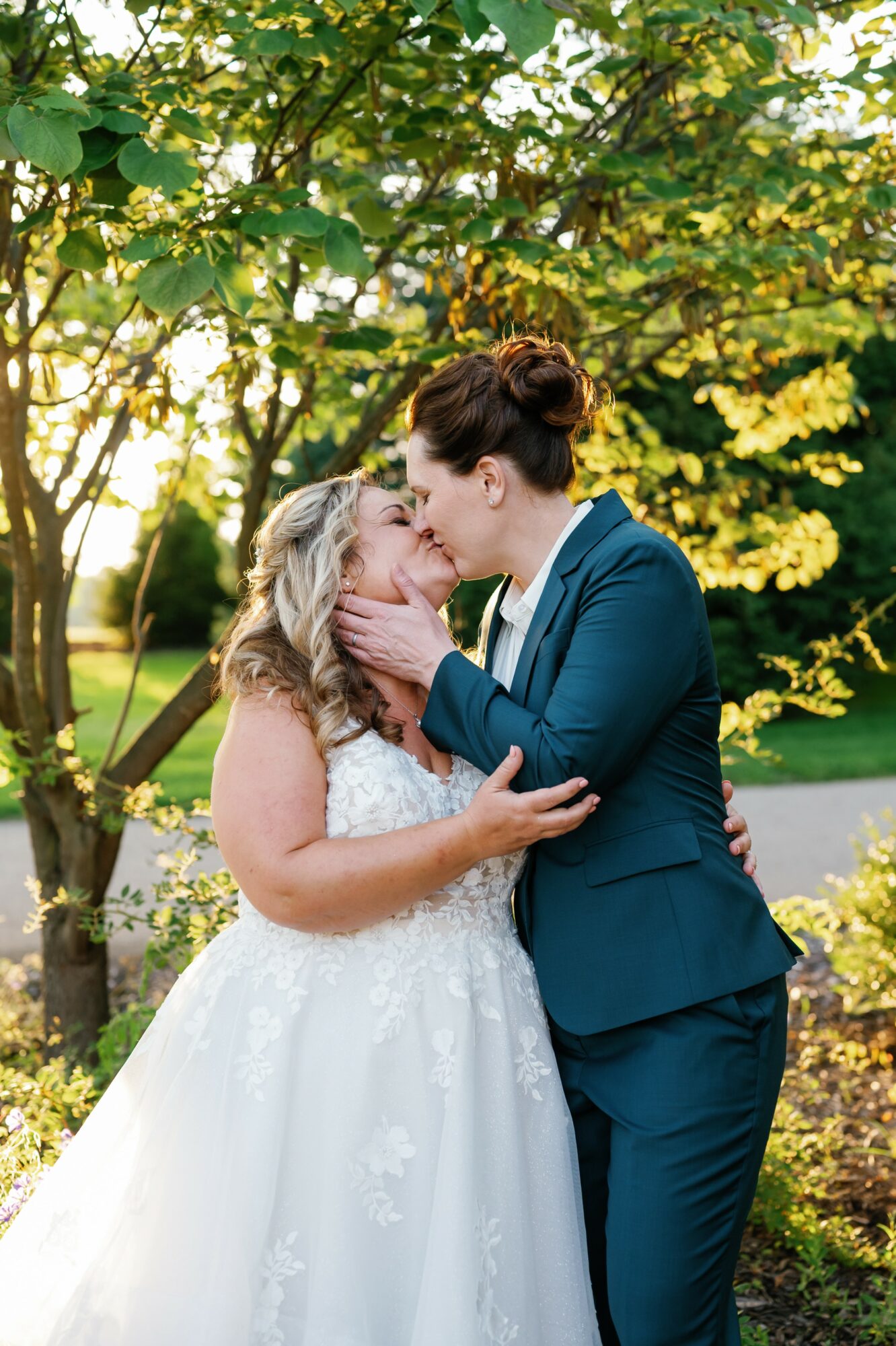 Two women, one in a wedding dress and the other in a suit, kiss outdoors with trees and sunlight in background.