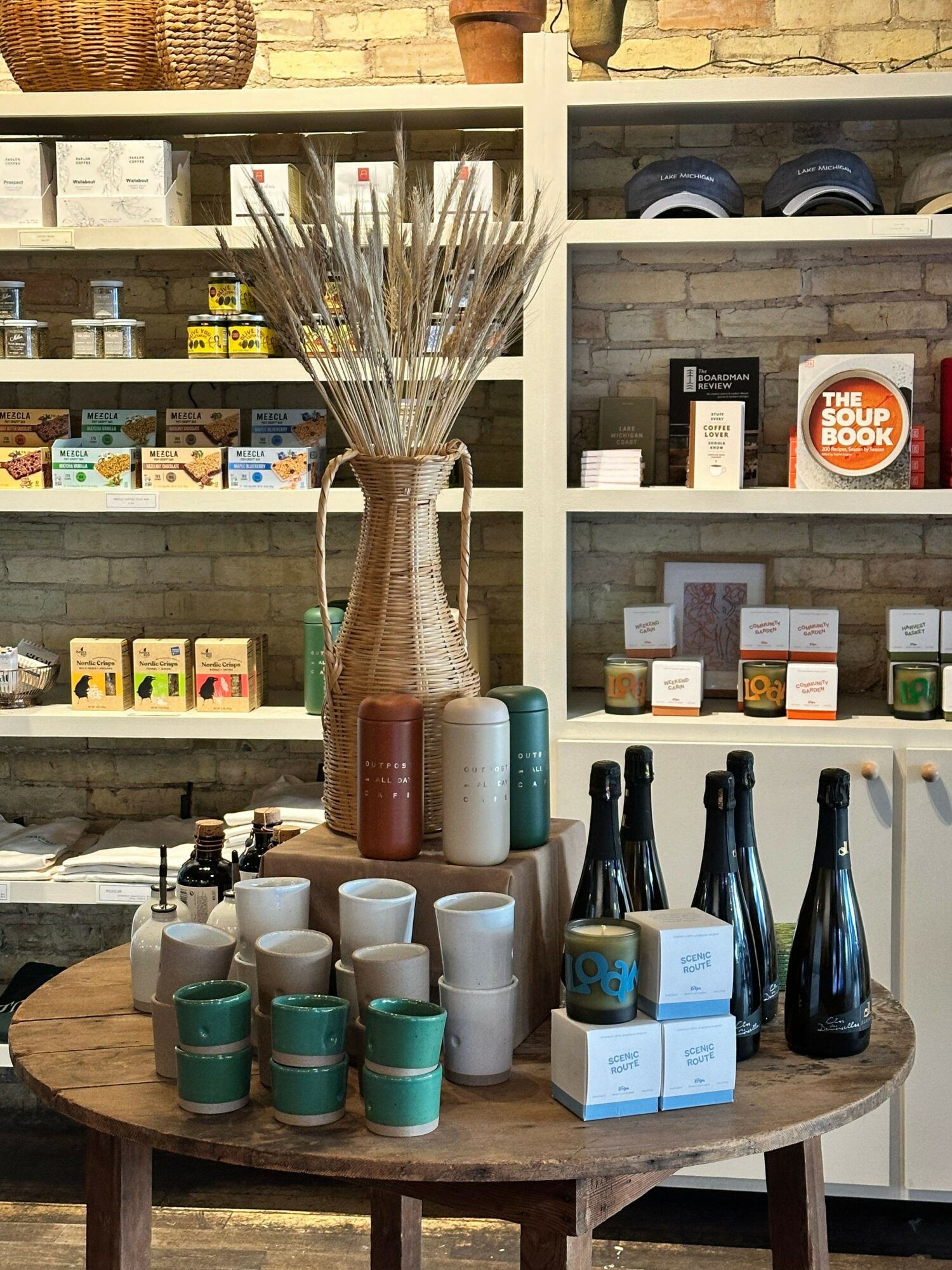 Display of bottles and cups on a round wooden table inside a shop, with shelves and products in the background.