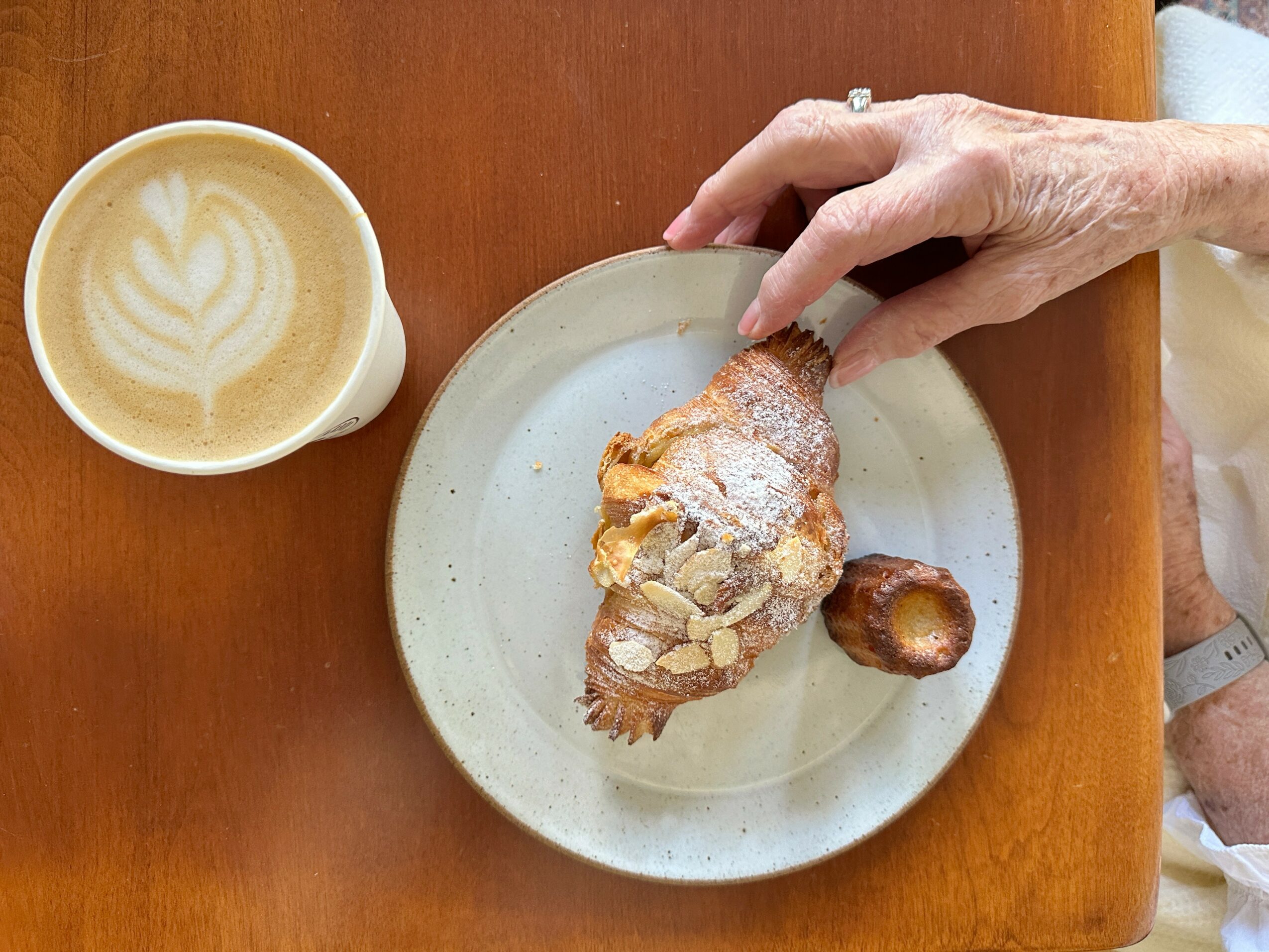 Hand reaching for a pastry on a plate, with a cup of latte with latte art on a wooden table.