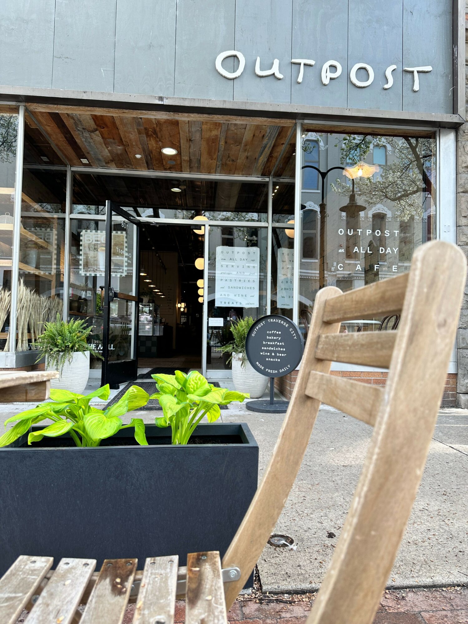 Outdoor seating area with a wooden chair and a table with green plants outside a cafe or shop.