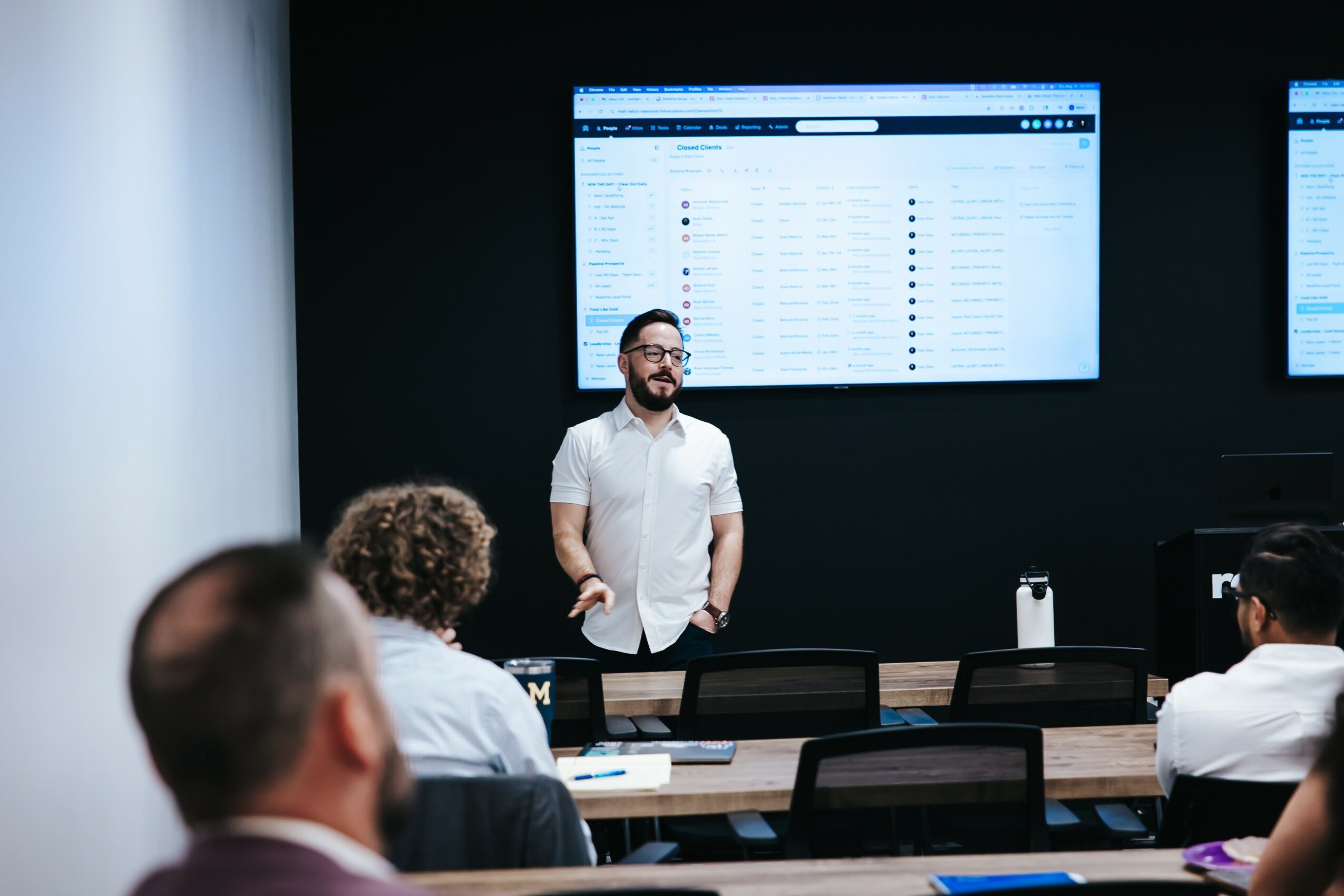 Person standing in front of a large screen displaying a computer interface, with seated audience members in a conference room.
