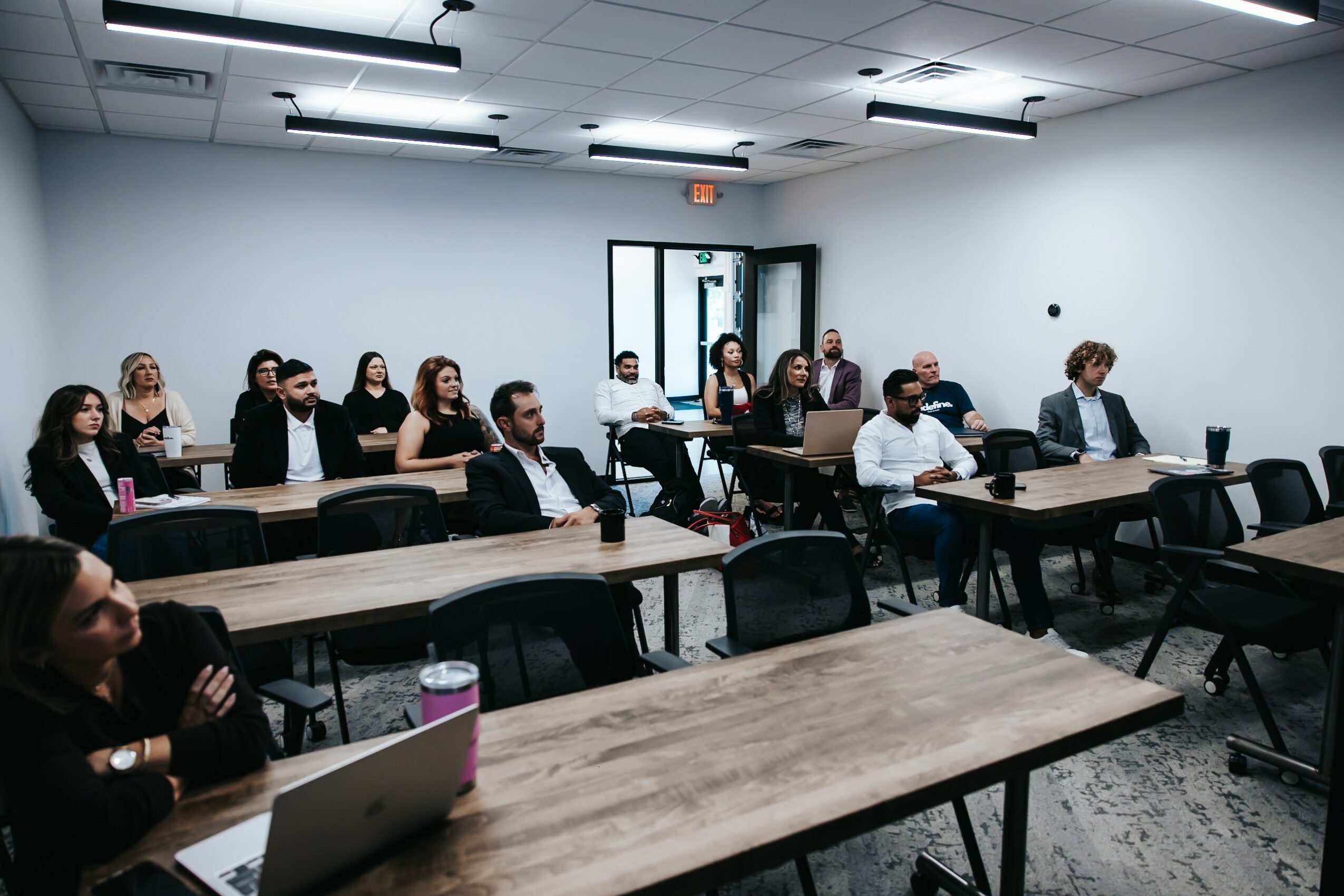 People seated in a classroom or conference room, facing forward, with tables and chairs, some using laptops.