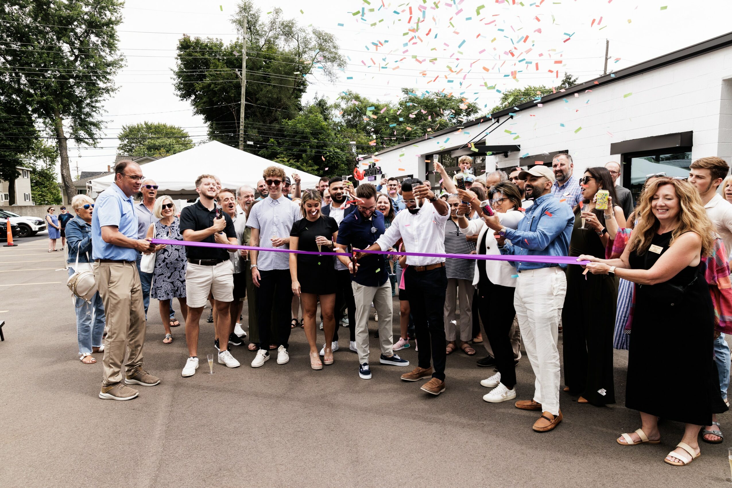 Group of people gathered outdoors for a ribbon-cutting event, some holding a pink ribbon, with tents and trees in background.