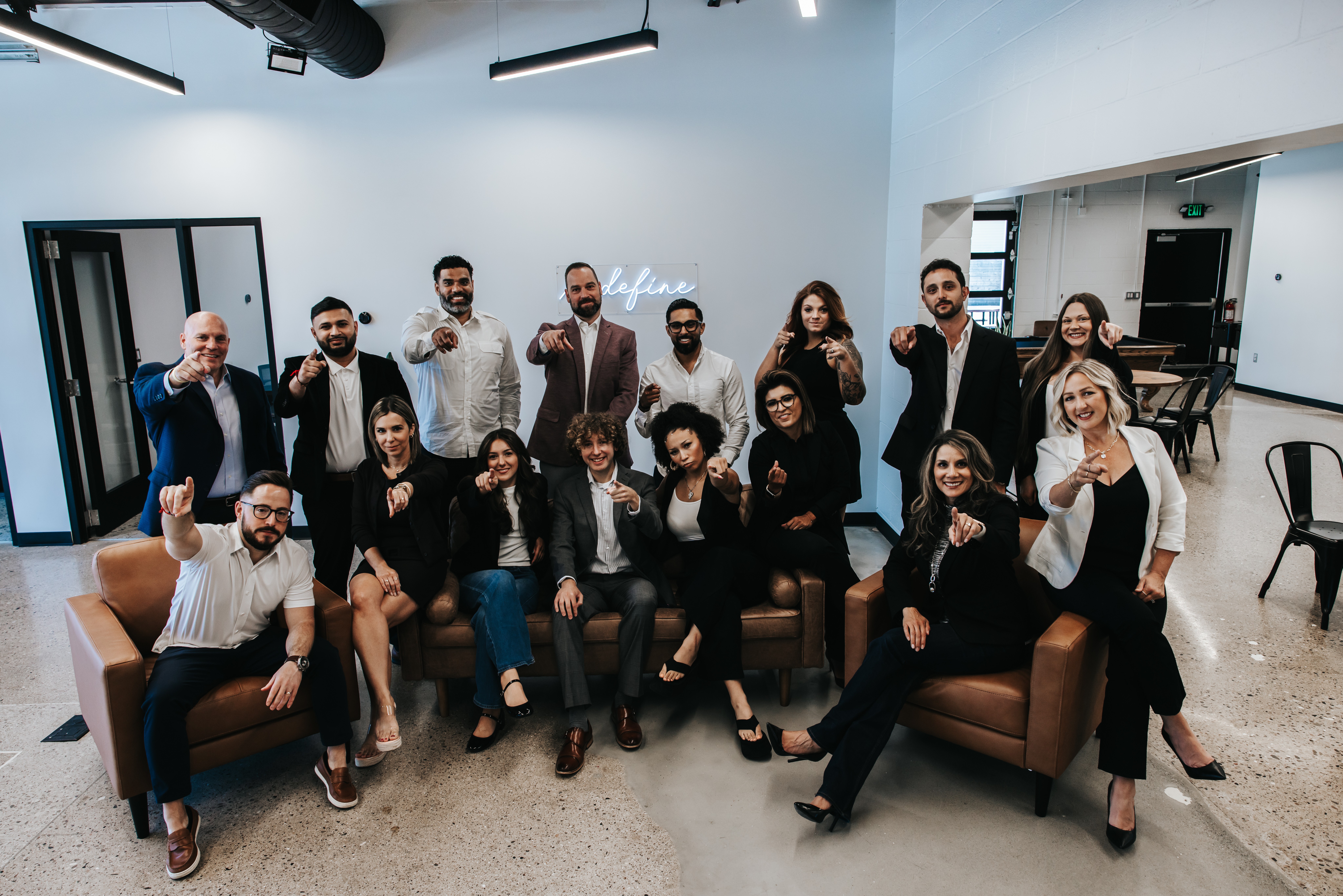 Group of 16 people in a modern office, some sitting on sofas and others standing, smiling and posing for the photo.