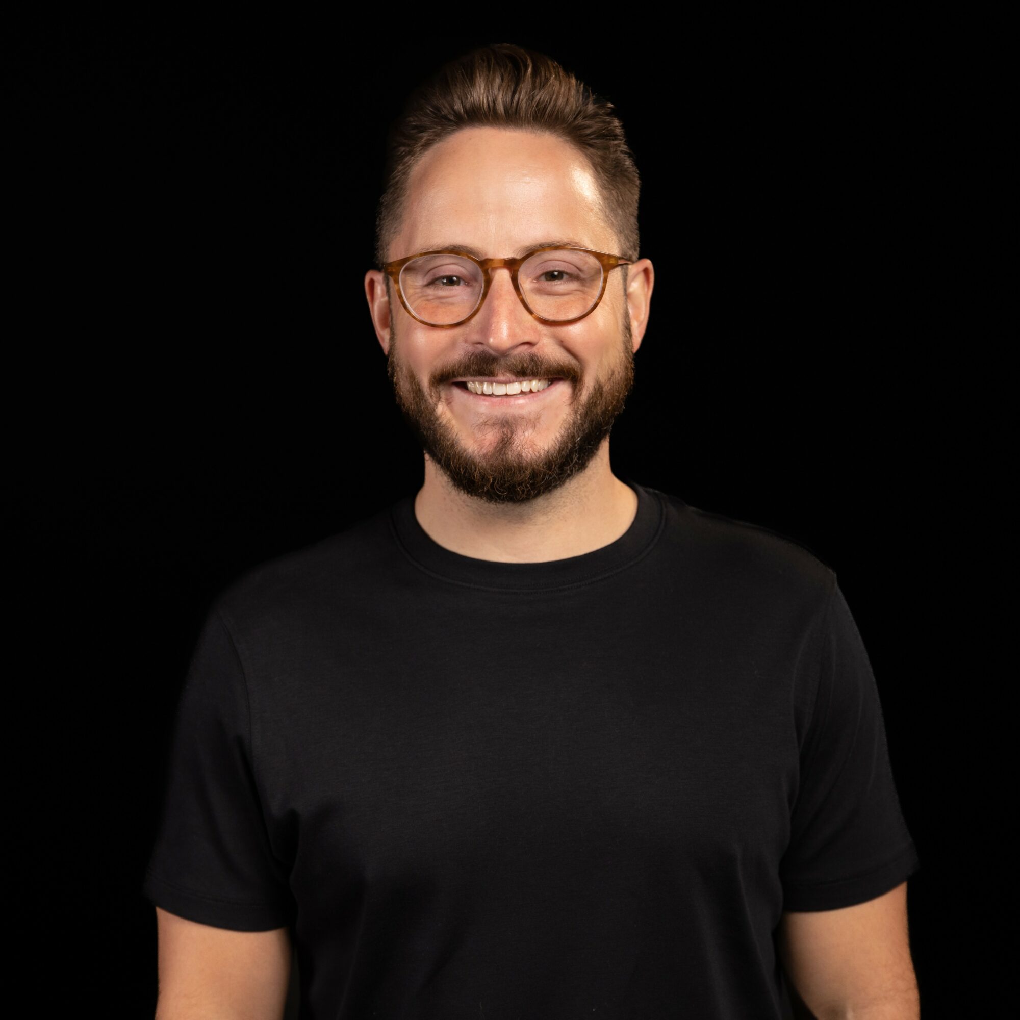 Smiling man with glasses and a beard, wearing a black t-shirt, against a black background.