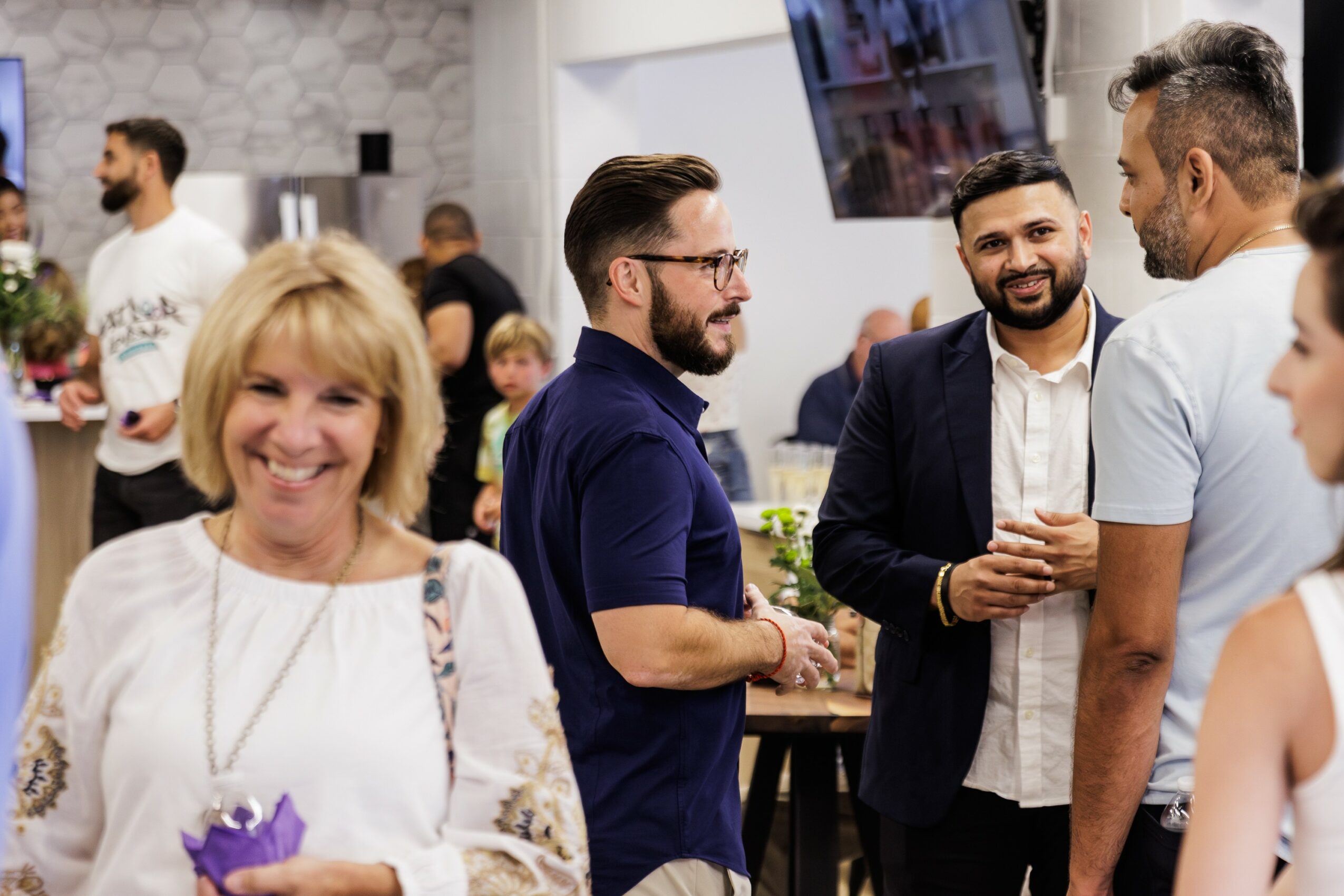 Group of people socializing at a gathering, smiling and talking, with a woman in the foreground and men in the background.