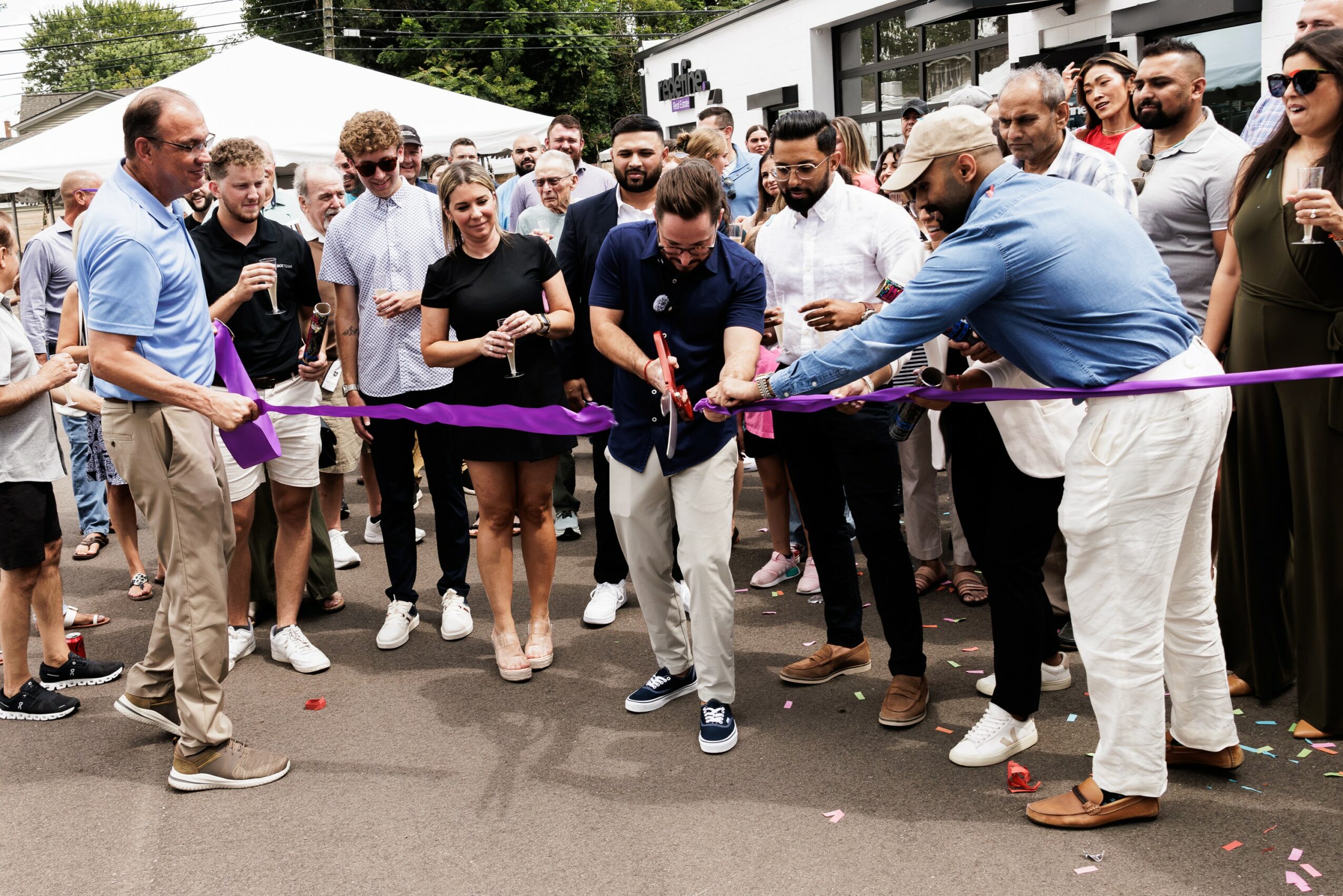 Group of people at a ribbon-cutting event outdoors, with a man cutting the ribbon and others watching, some holding drinks.