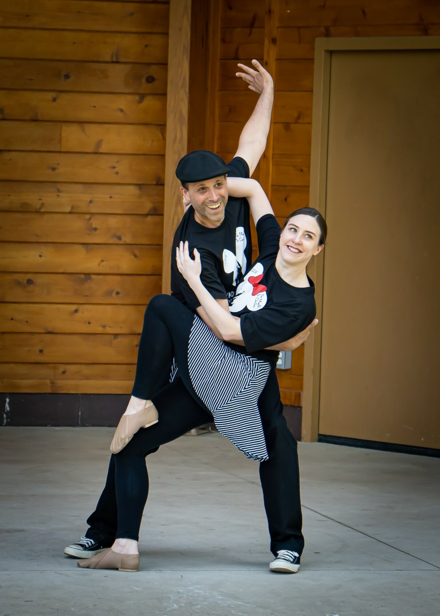 Two dancers perform a lift indoors, with wooden wall background, smiling and looking at each other, one wearing a black hat.