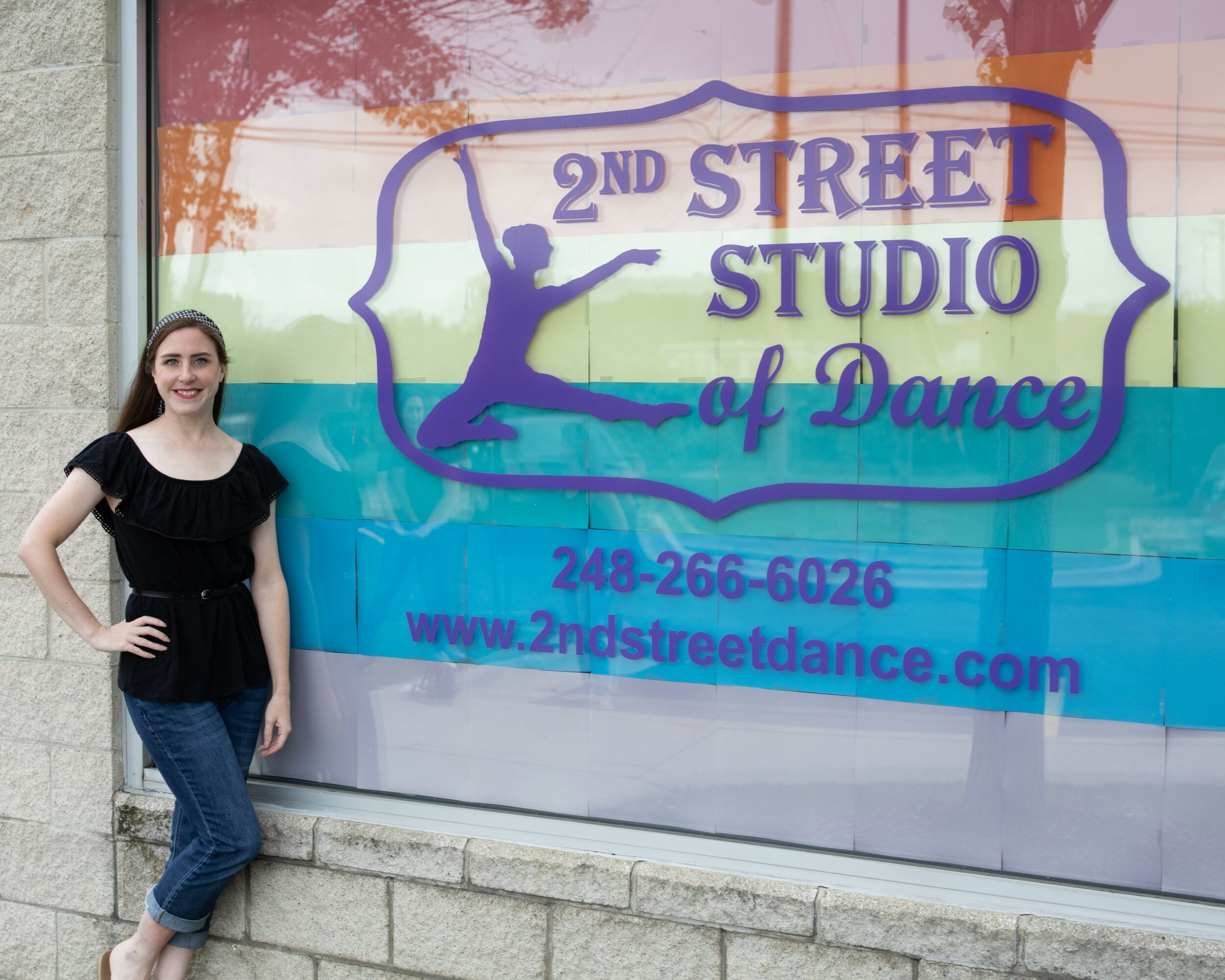 Woman standing outside a dance studio window with colorful sign, smiling, wearing black top and jeans.