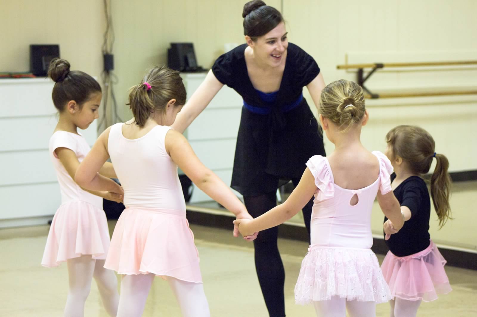 Dance instructor with five young girls holding hands in a dance studio, all wearing ballet outfits.