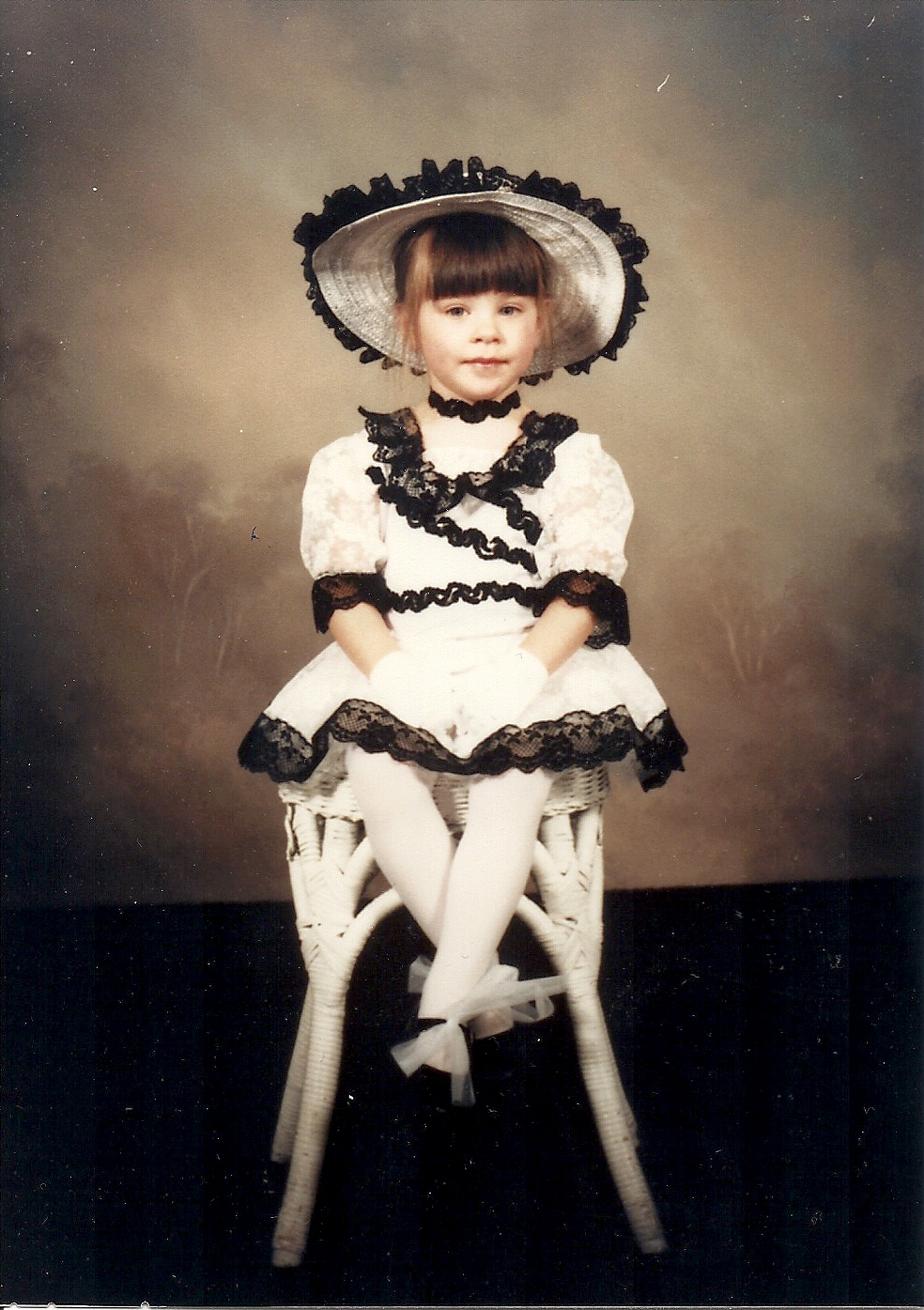 Child in vintage dress and hat sitting on a chair, with a neutral background.