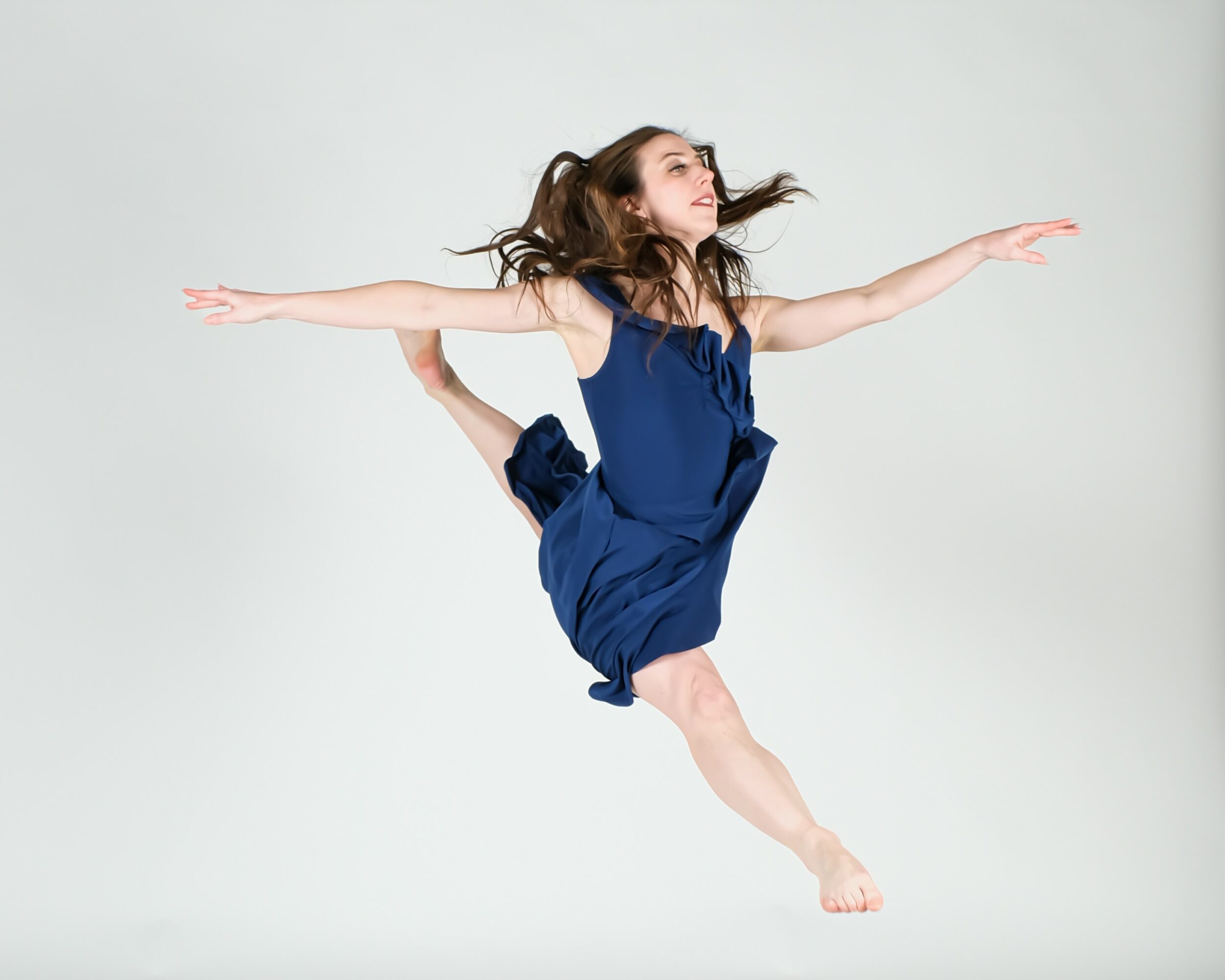 Young woman in a blue dress performs a dance leap with arms extended and hair flowing, against a plain background.