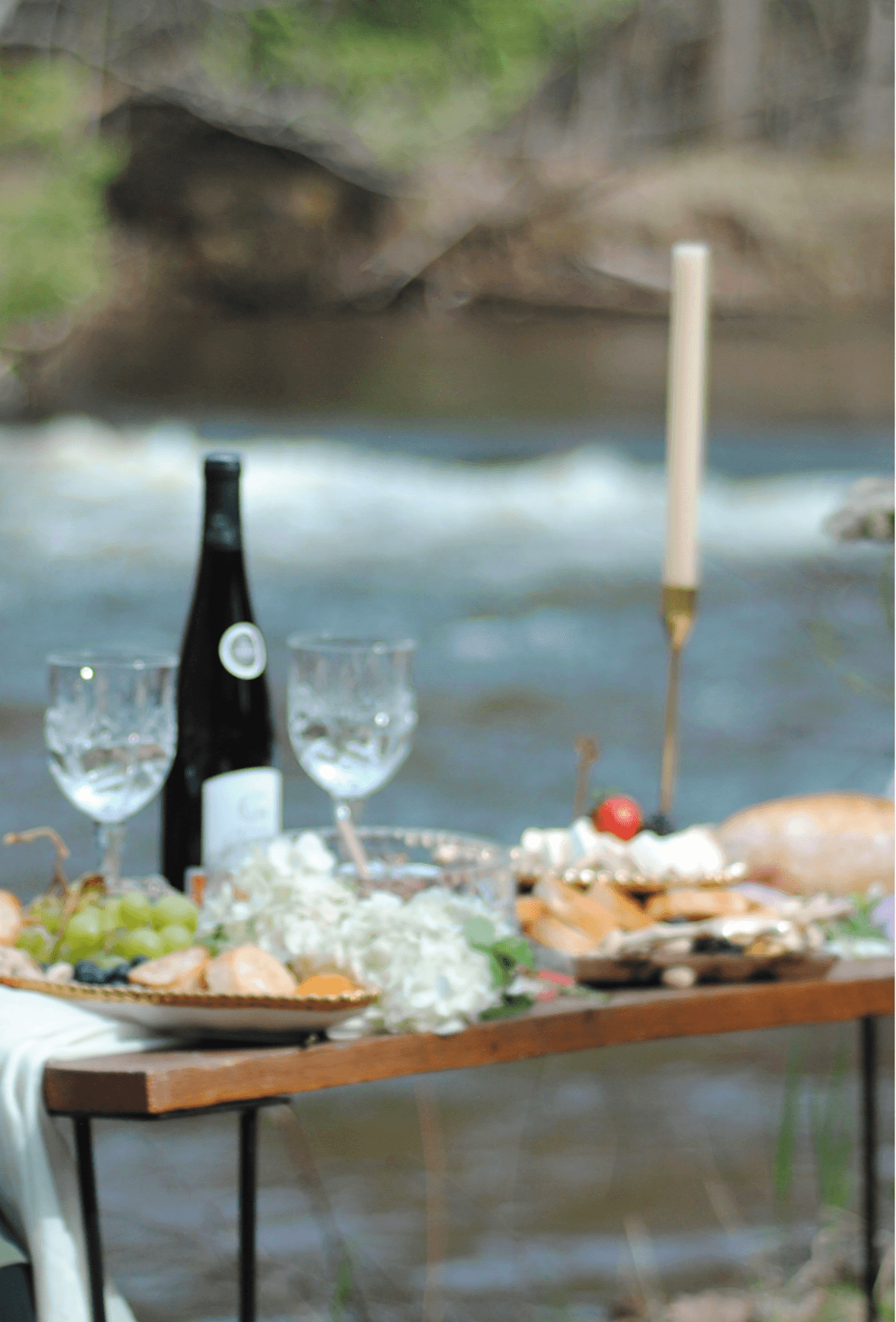 Picnic table set with wine, glasses, food, and a bottle near a river with a candle and rocks in the background.