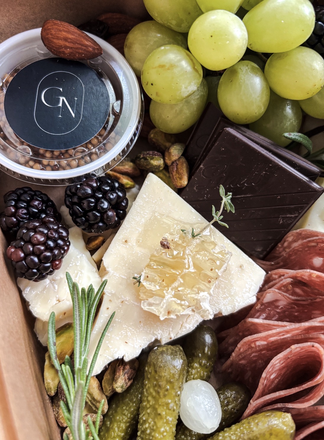 Assorted cheeses, green grapes, blackberries, cured meats, chocolate, nuts, and a small container of honey or jam.