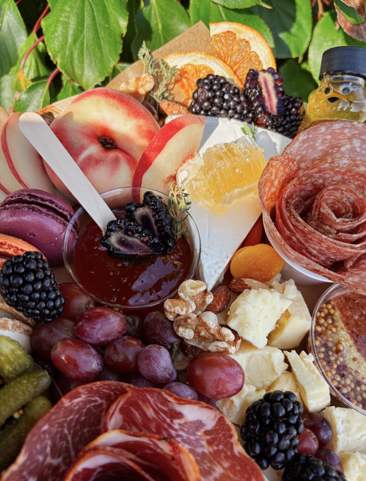 Assorted fresh fruits, jams, and cheeses arranged with leaves and flowers, including peaches, grapes, blackberries, and orange slices.