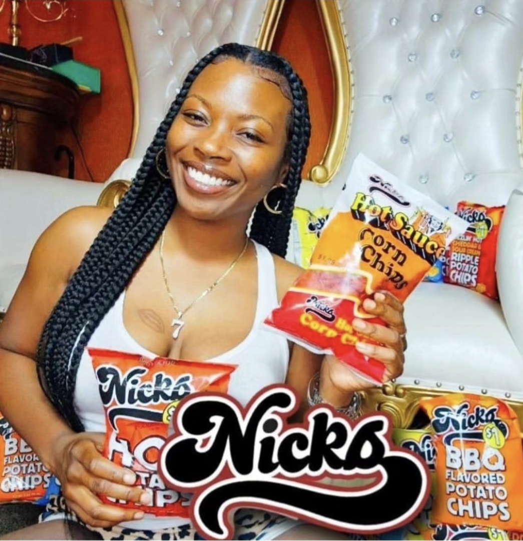 Smiling woman with braided hair holding a snack bag, sitting in a room with a white tufted headboard and snack packages behind her.