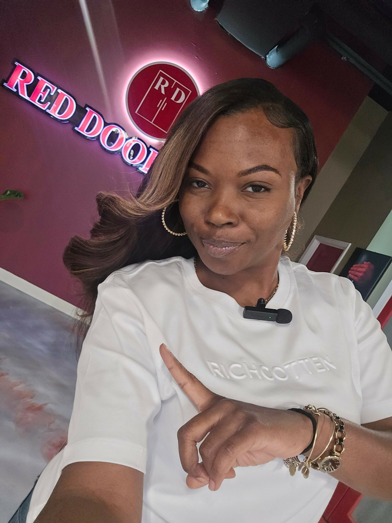 Woman with long hair wearing a white shirt, pointing upward, in front of a red and pink sign that says 'Red Door'.