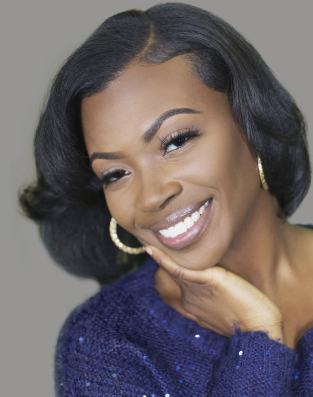 Smiling woman with short black hair, wearing gold hoop earrings and a blue sweater, resting her chin on her hand.