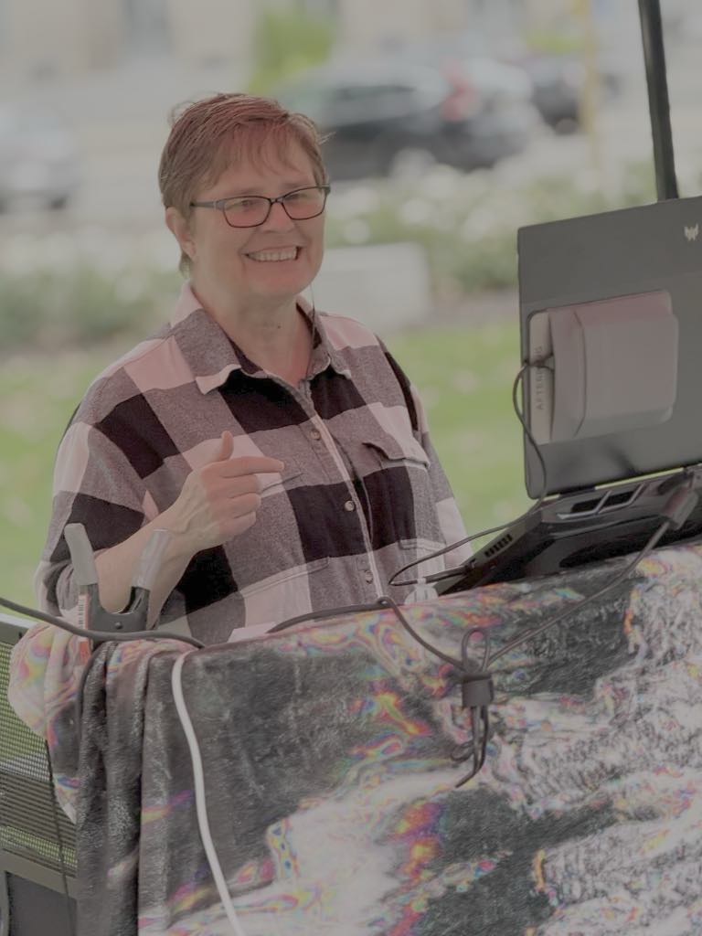 Smiling woman with short hair and glasses giving a thumbs-up, standing behind a counter with a computer, in bright light.