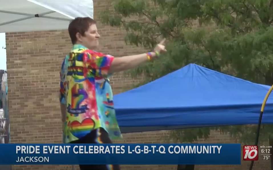 Person with short hair wearing a colorful shirt, standing outdoors near tents, celebrating at pride event.