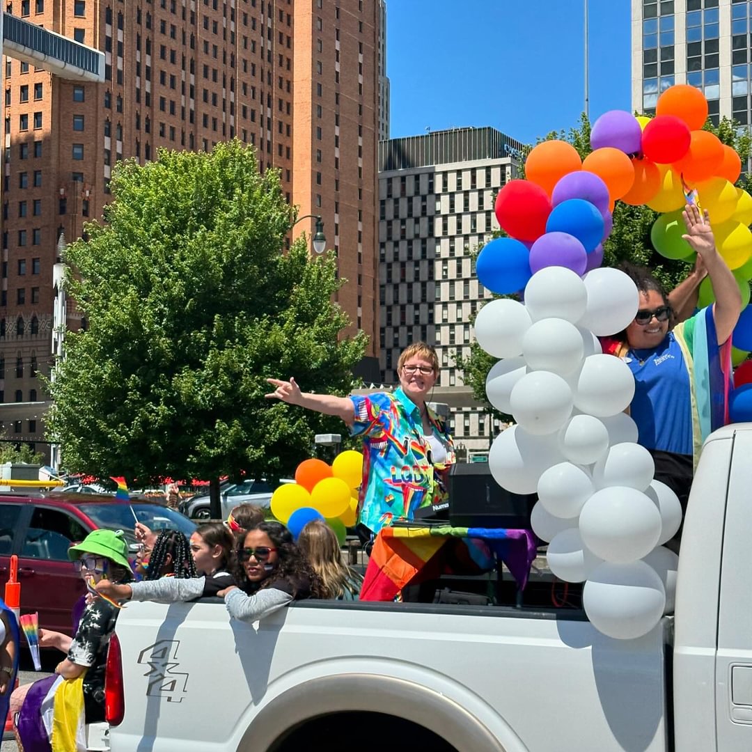 People celebrating on a truck with colorful balloons in an urban setting, smiling and waving, with tall buildings and a tree in the background.