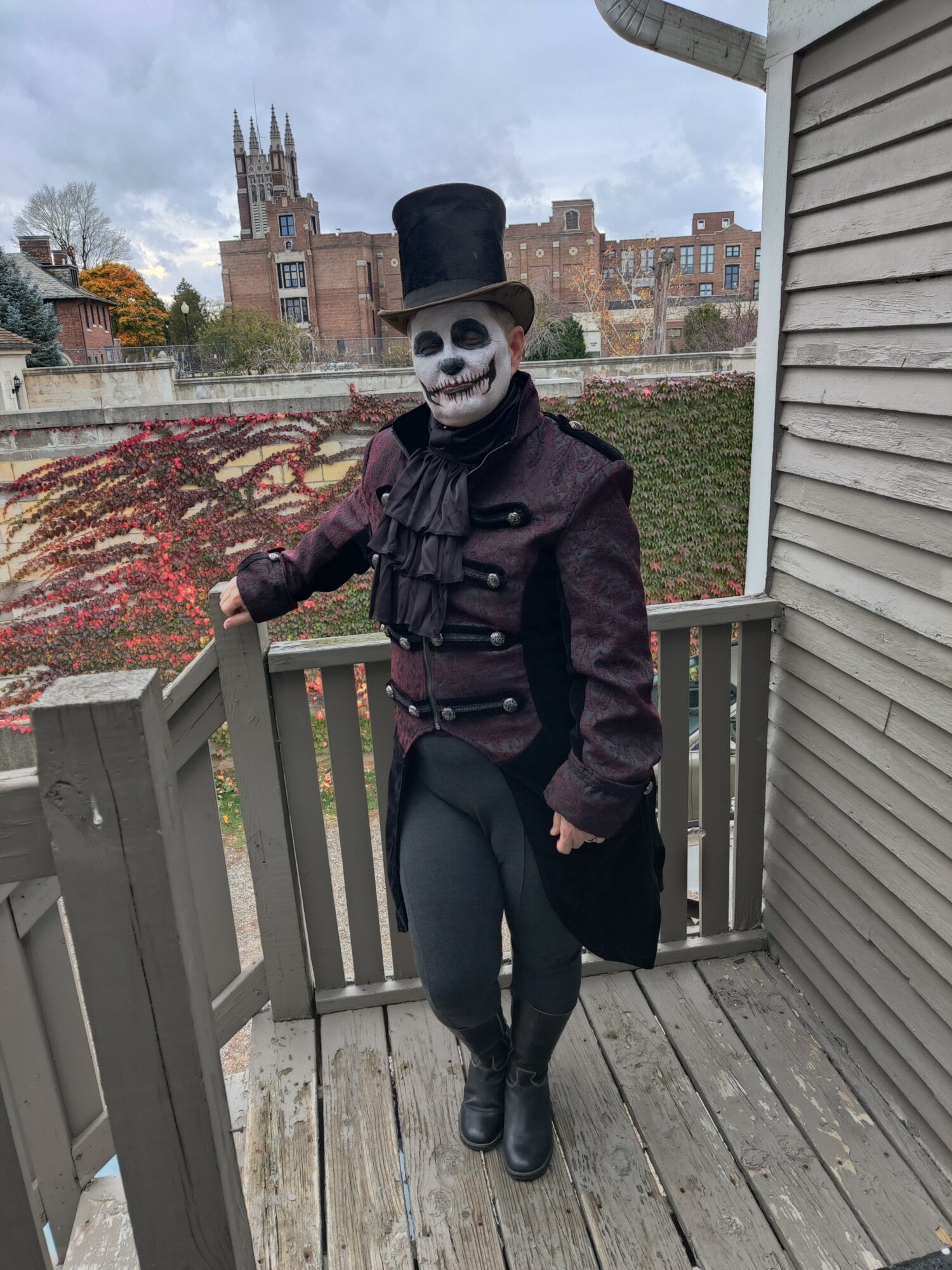 Person dressed as a skeleton with a top hat and dark clothing standing on a wooden porch railing, with a church and trees in the background.