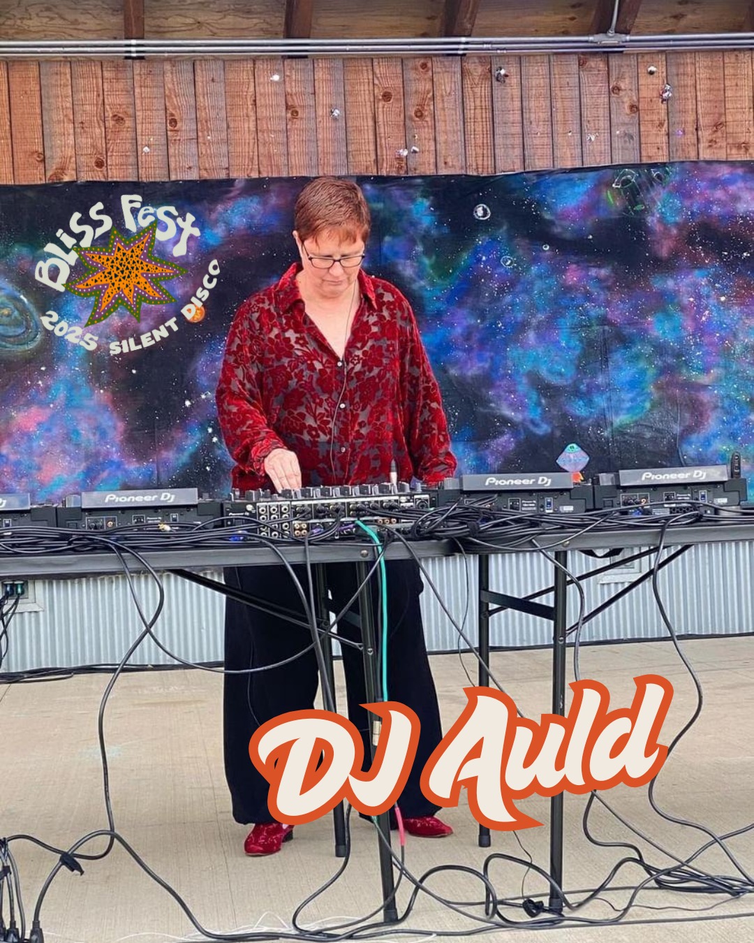Woman DJ wearing red patterned top and glasses, standing behind DJ equipment outdoors, with a colorful galaxy backdrop and a sign reading 'Bliss Fest 2023 Silent Disco'.