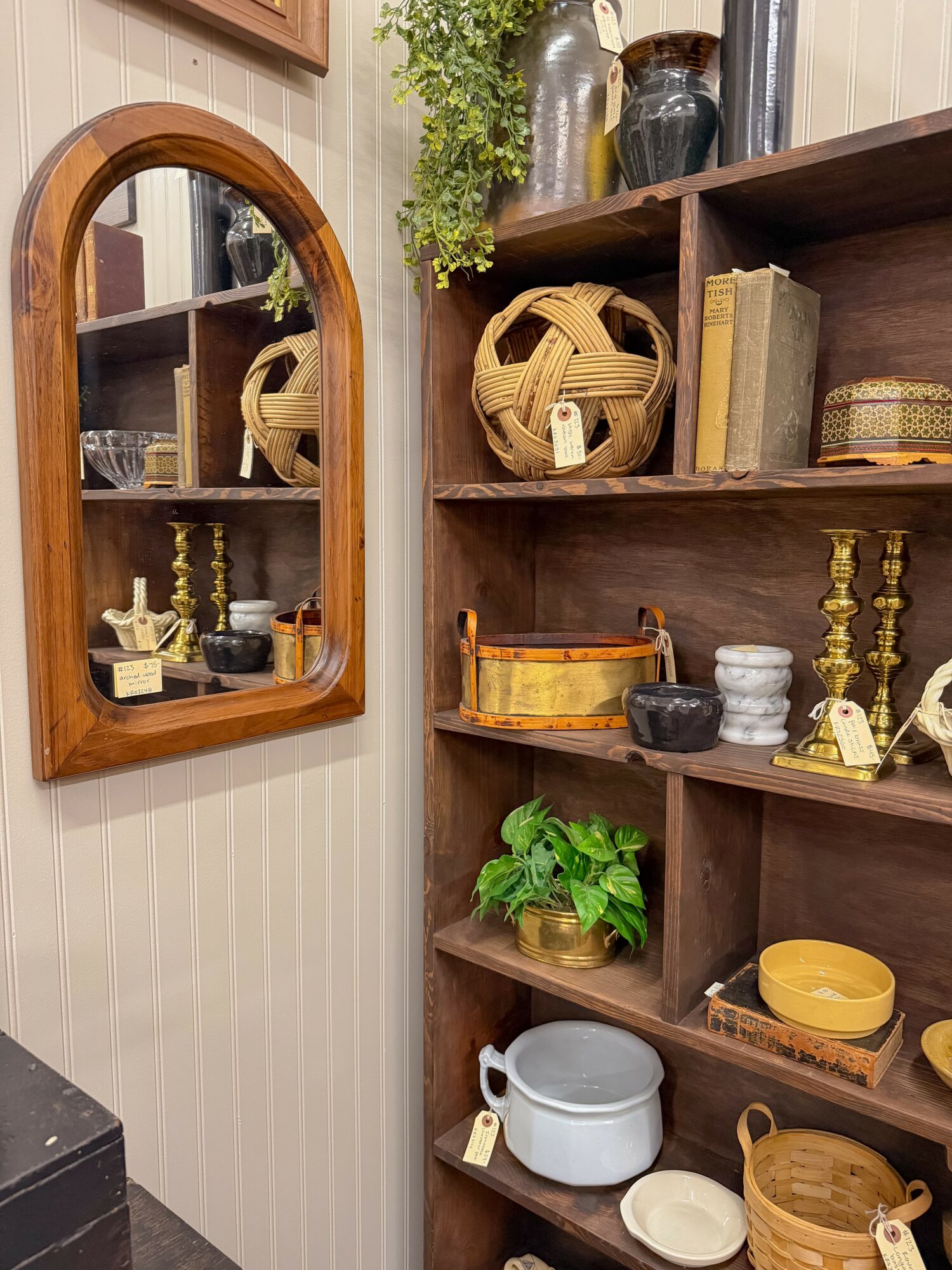 Wooden shelf with decorative items, books, and plants, reflected in an arched mirror on a wall with vertical striped wallpaper.