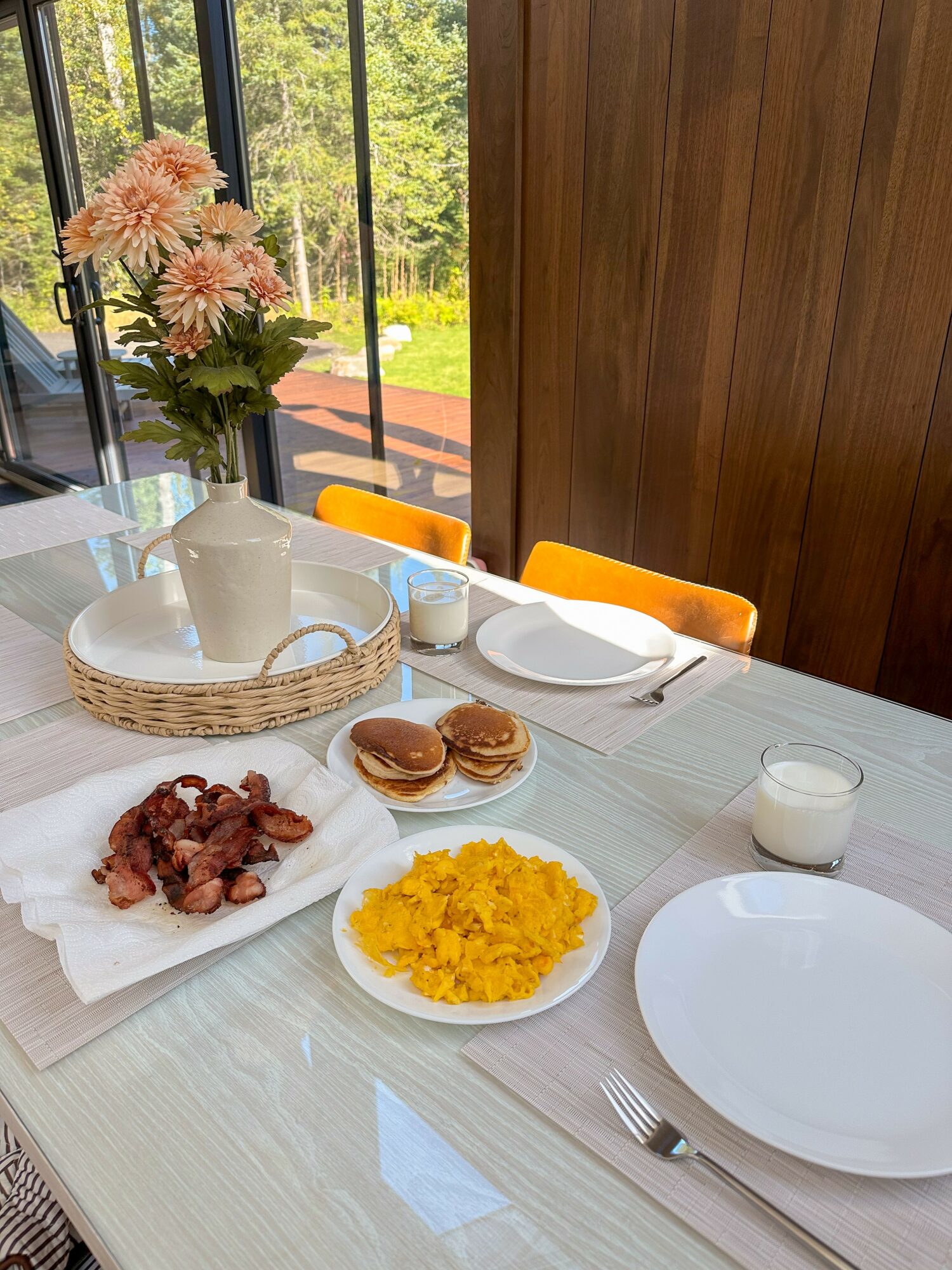 Table with flowers, breakfast items including bacon, pancakes, scrambled eggs, milk, and empty plates, set near a window with outdoor view.