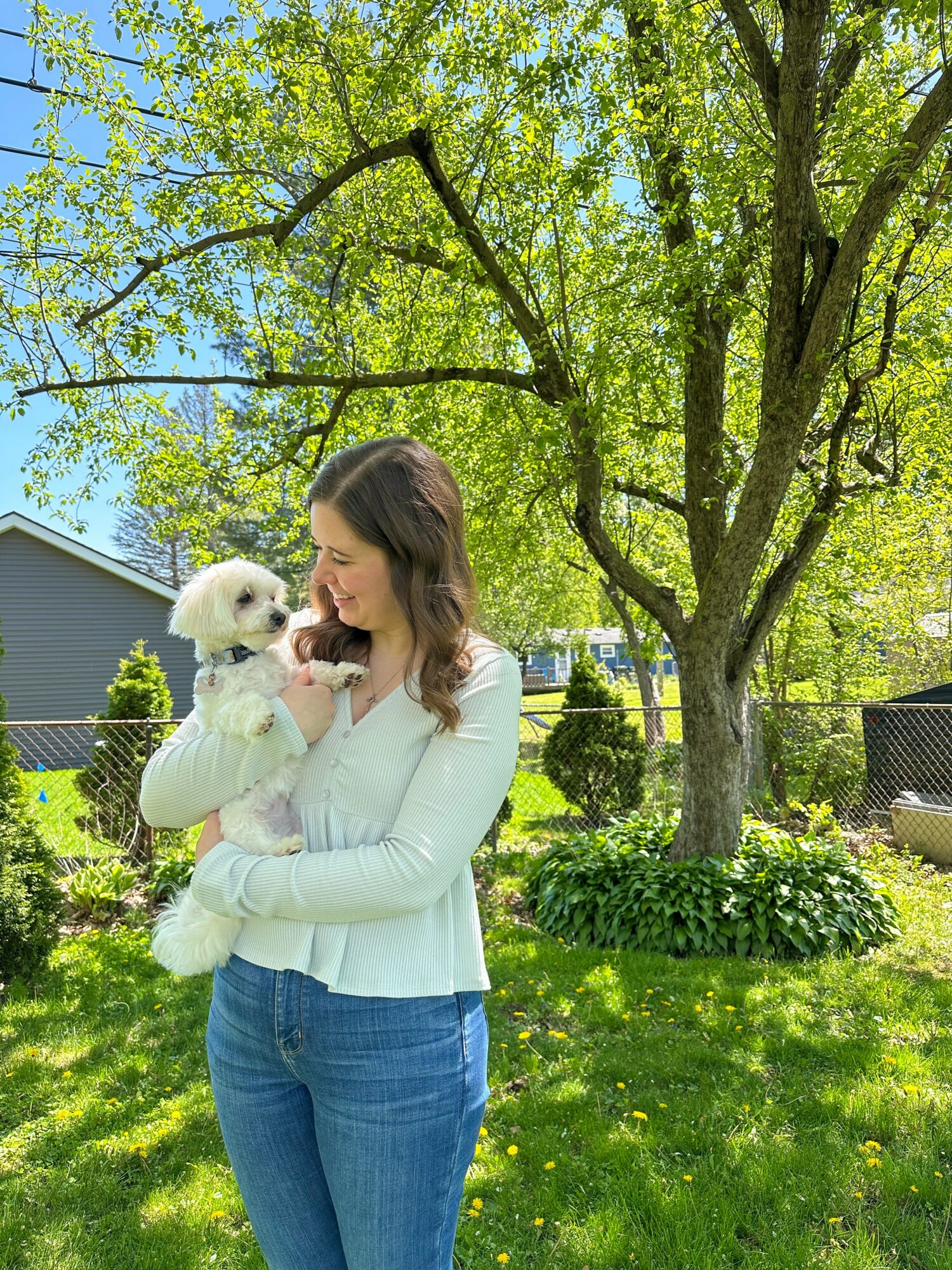 Woman holding a small white dog outdoors in a green yard with trees and a house in the background.