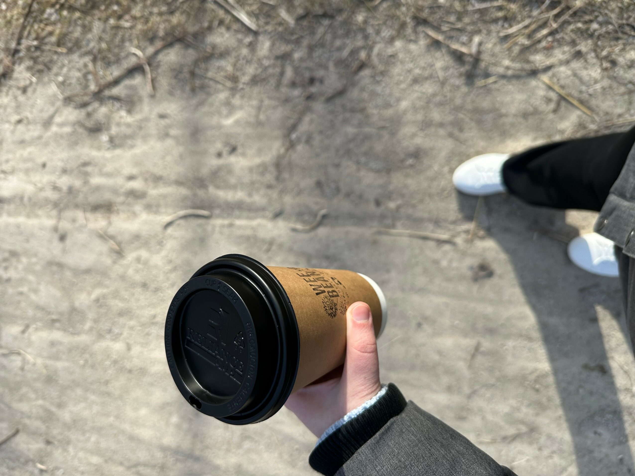 Person holding a coffee cup with a black lid, wearing a gray sleeve, on a concrete surface.