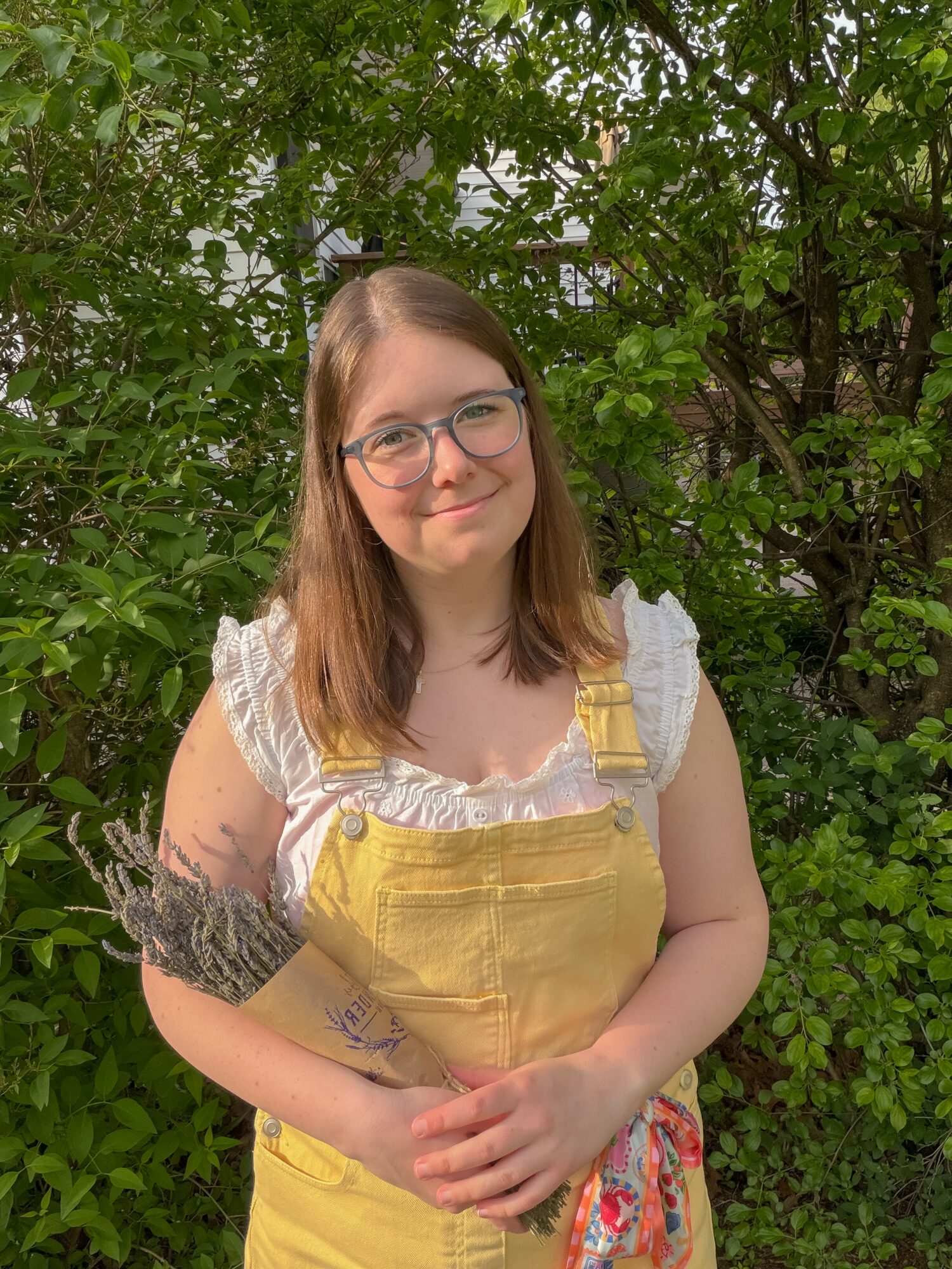 Young woman with glasses and shoulder-length hair standing outdoors in front of green foliage, holding a lavender bouquet.