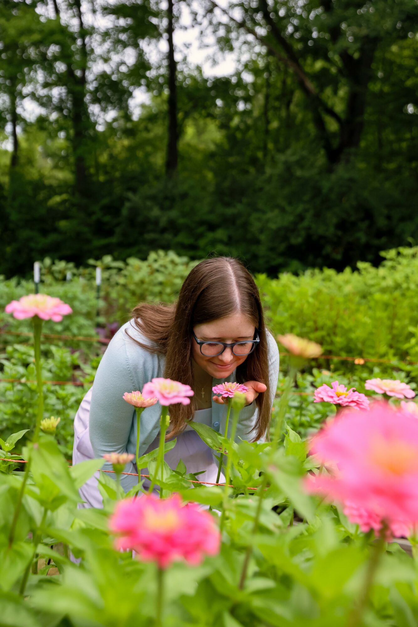 Woman with glasses leaning over pink flowers in a garden, surrounded by green plants and trees.