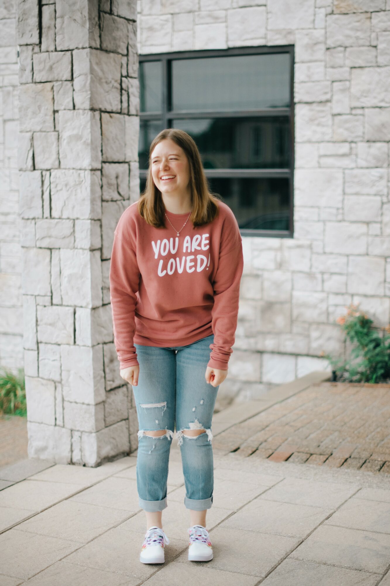 Young woman with shoulder-length hair smiling, wearing a red sweatshirt with white text, ripped jeans, and white sneakers, standing outside near a stone wall and window.