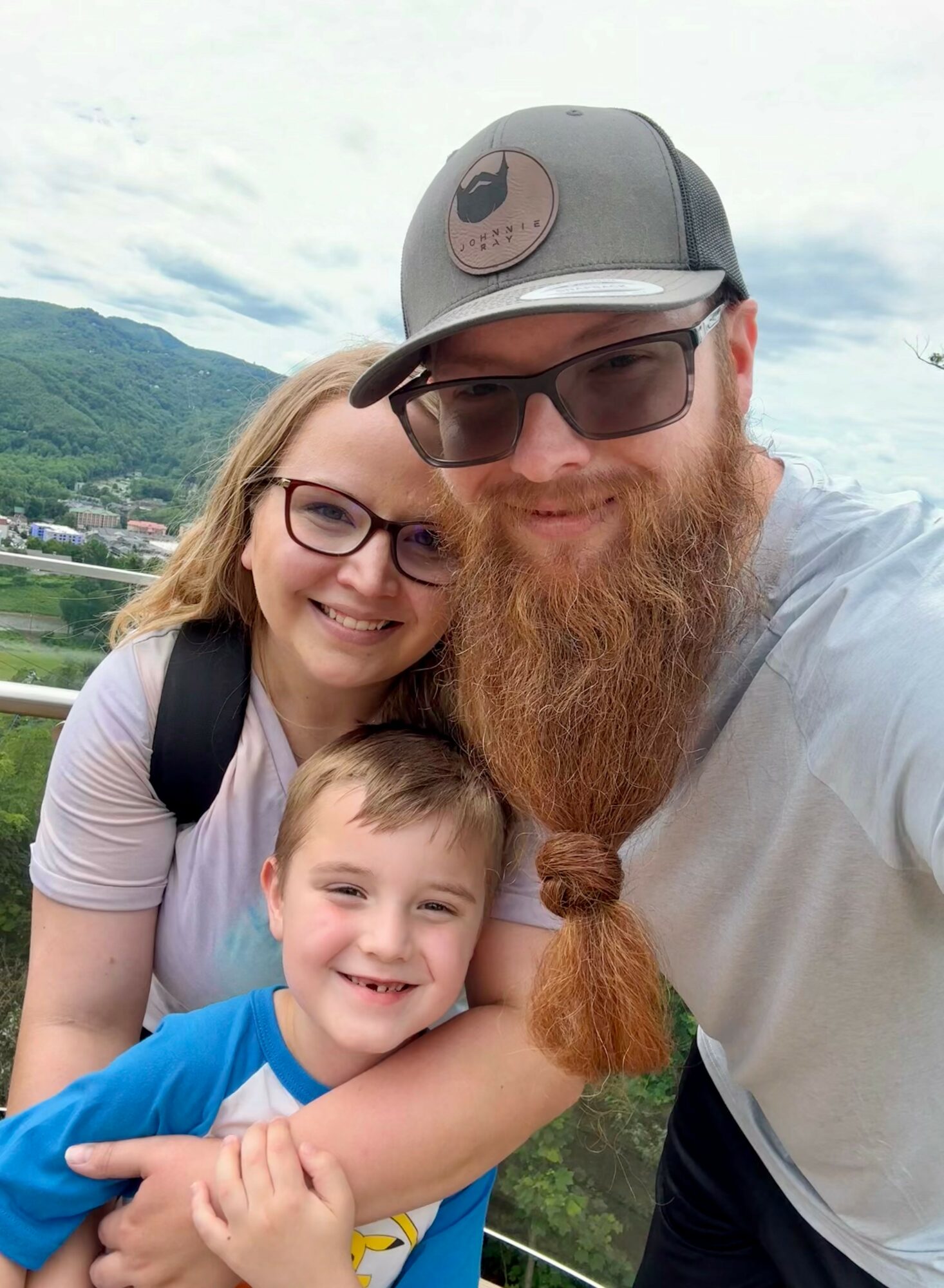 A smiling family of three outdoors with a scenic background, including a woman, a man with a beard, and a young boy.