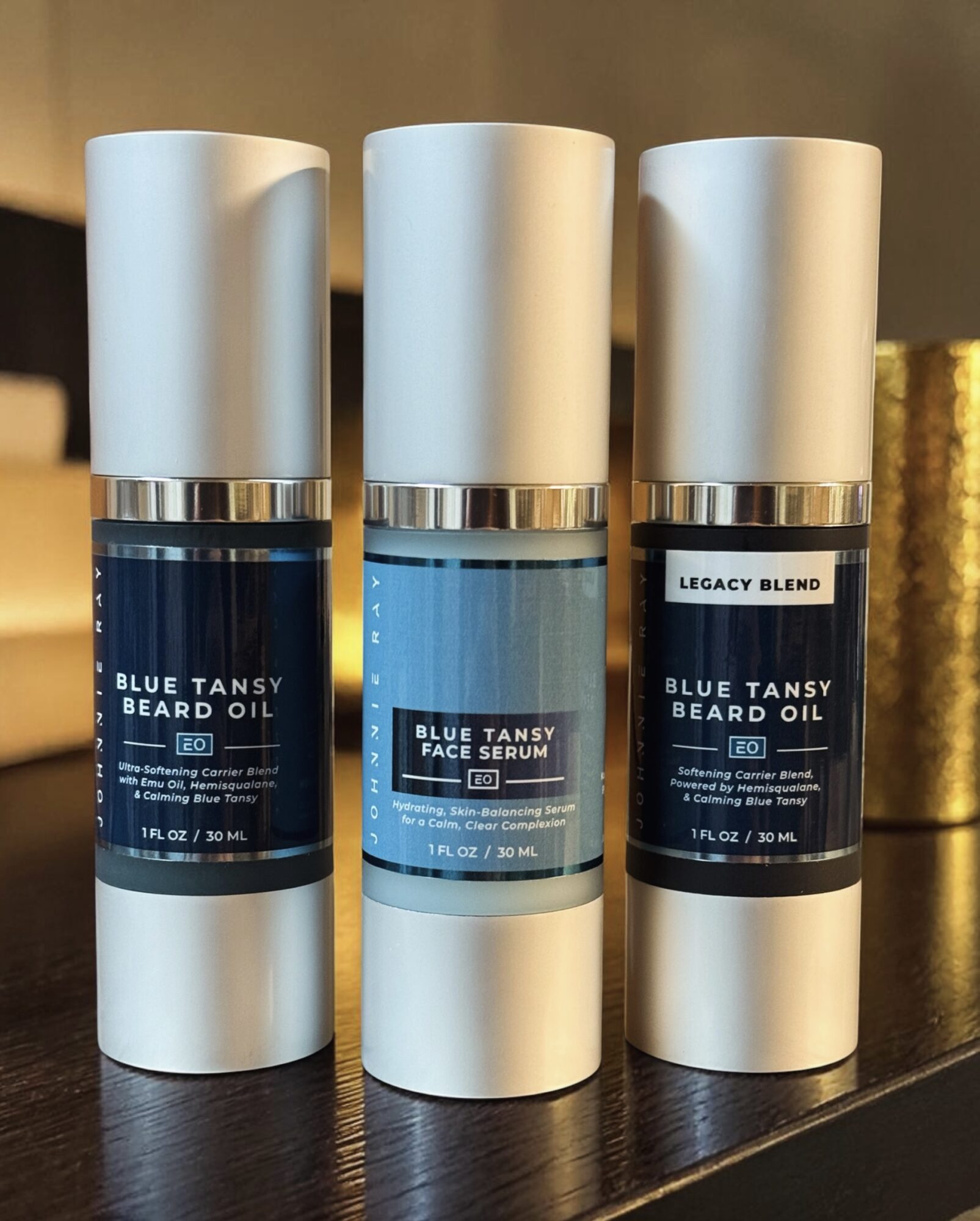 Three white bottles with blue labels and silver caps on a surface, containing beard oil and serum products.