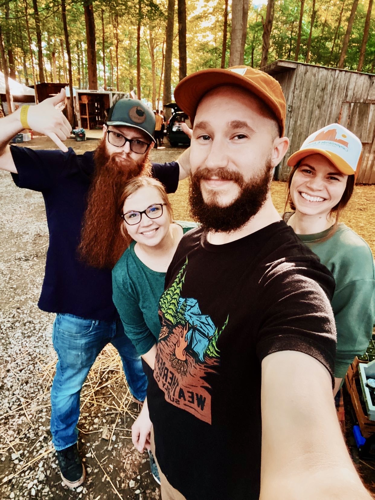 Four people smiling outdoors in a wooded area, taking a selfie together, with trees and a wooden structure in the background.