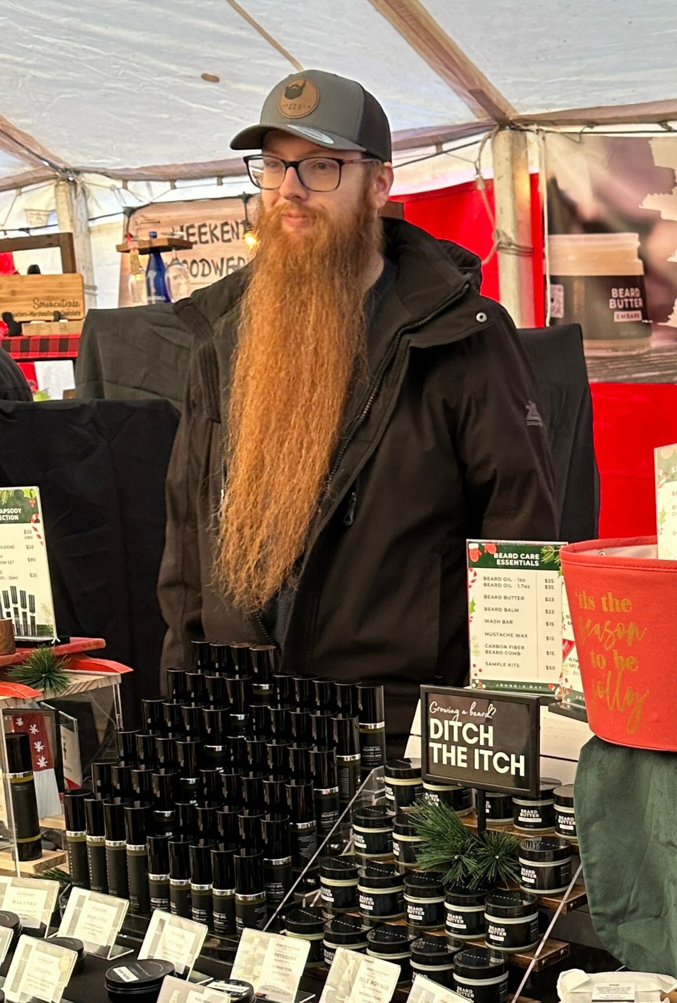 Man with long beard, glasses, black jacket, and cap standing behind a booth with small bottles and signs.